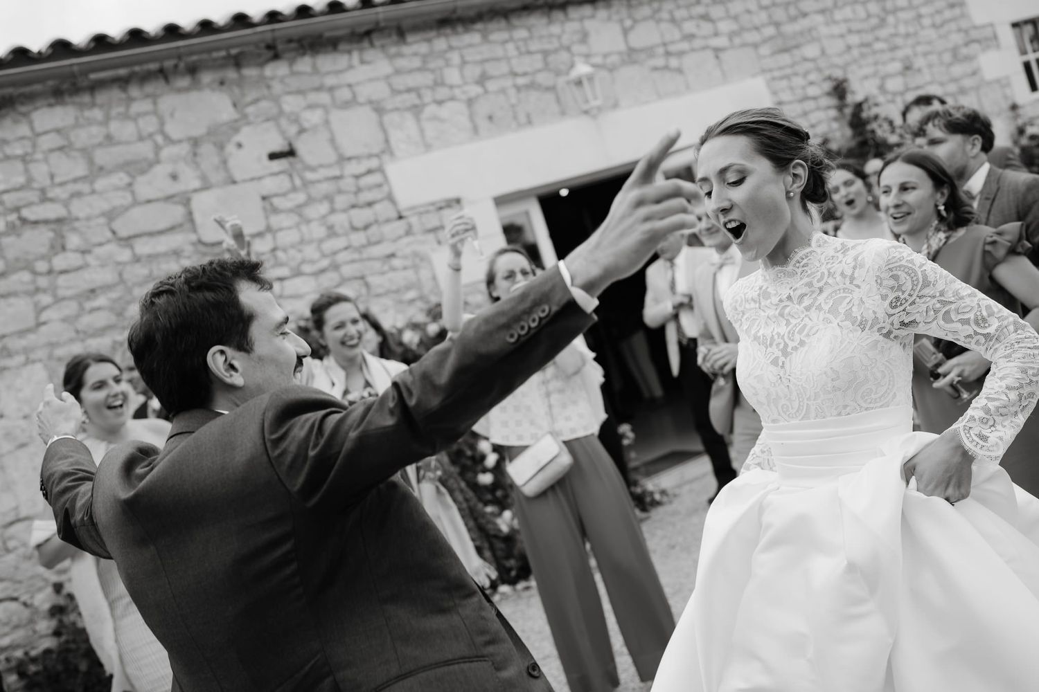 Wedding guests dancing and celebrating joyfully at an outdoor reception with stone walls in the background.