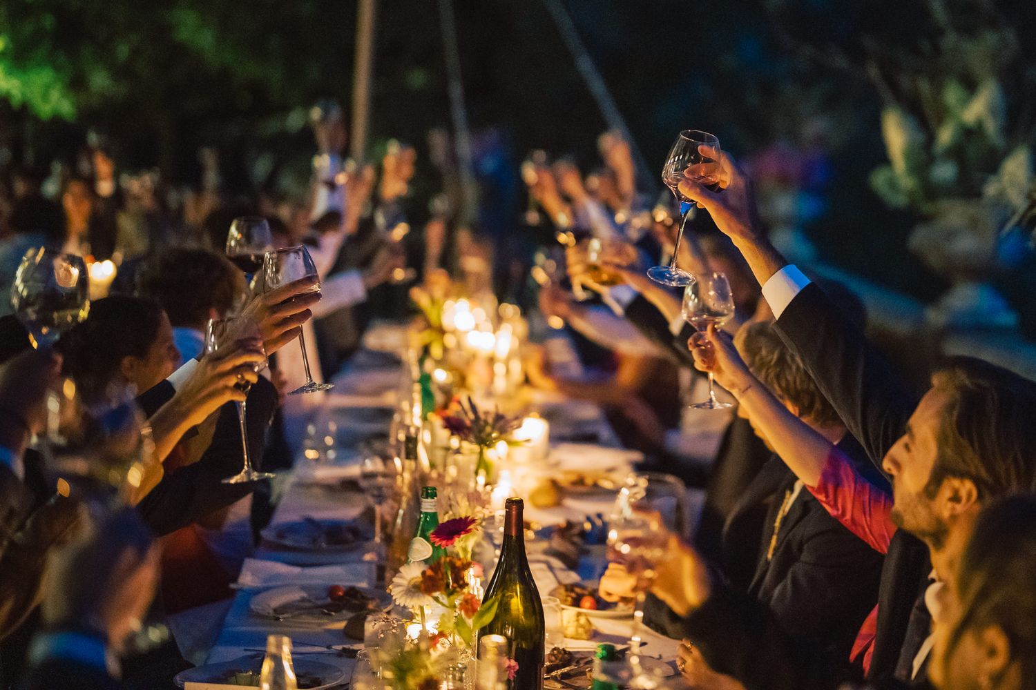 Candlelit dinner tables at dusk with guests celebrating and raising glasses.