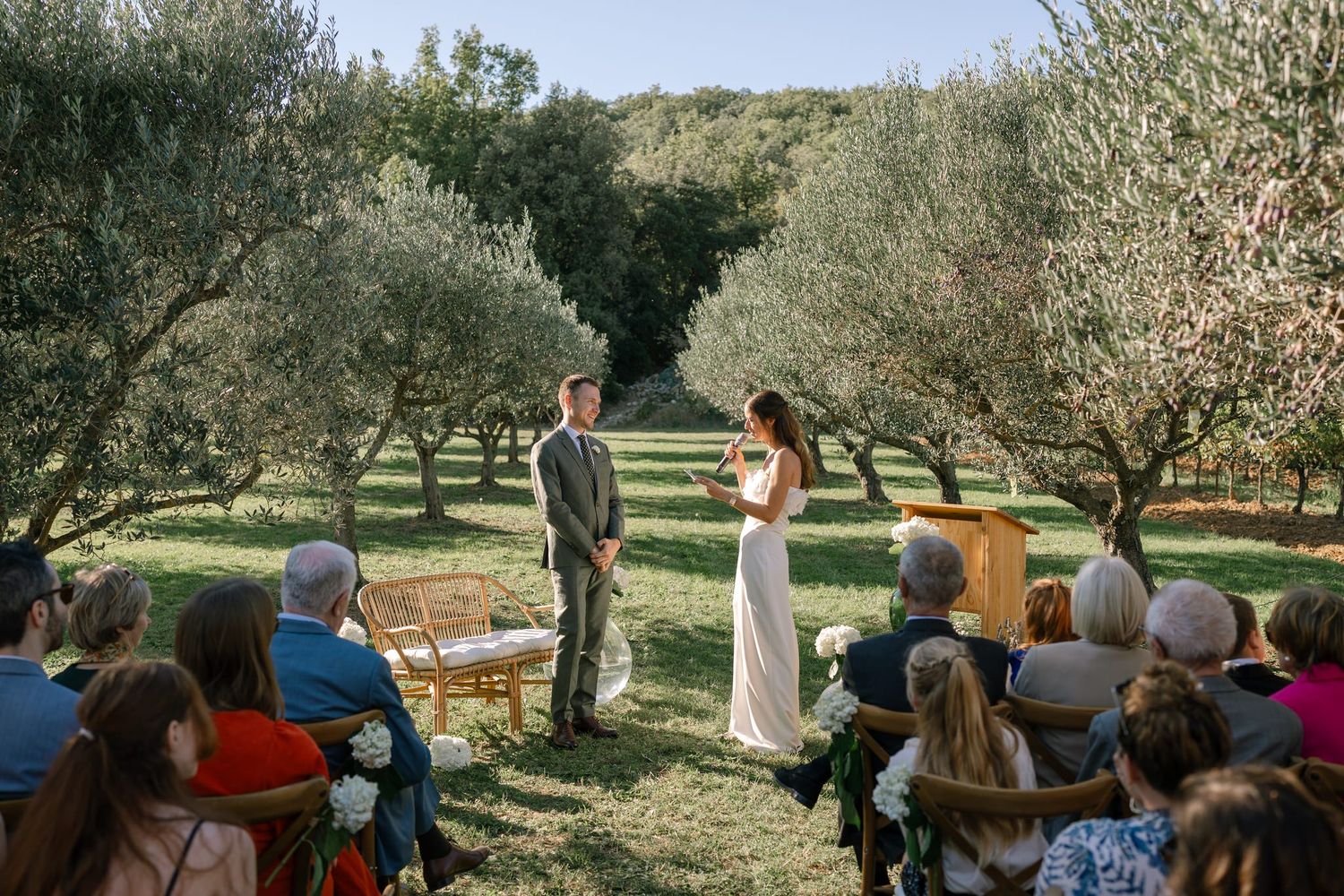 Wedding ceremony unfolds beneath olive trees with guests watching from white chairs.