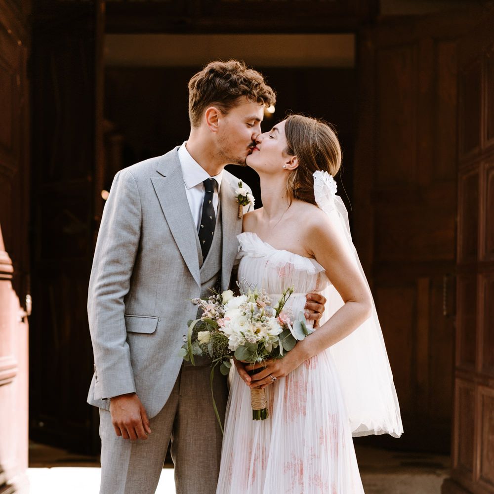 A romantic wedding moment with a couple in a grey suit and white dress holding a bouquet outside a wooden building.