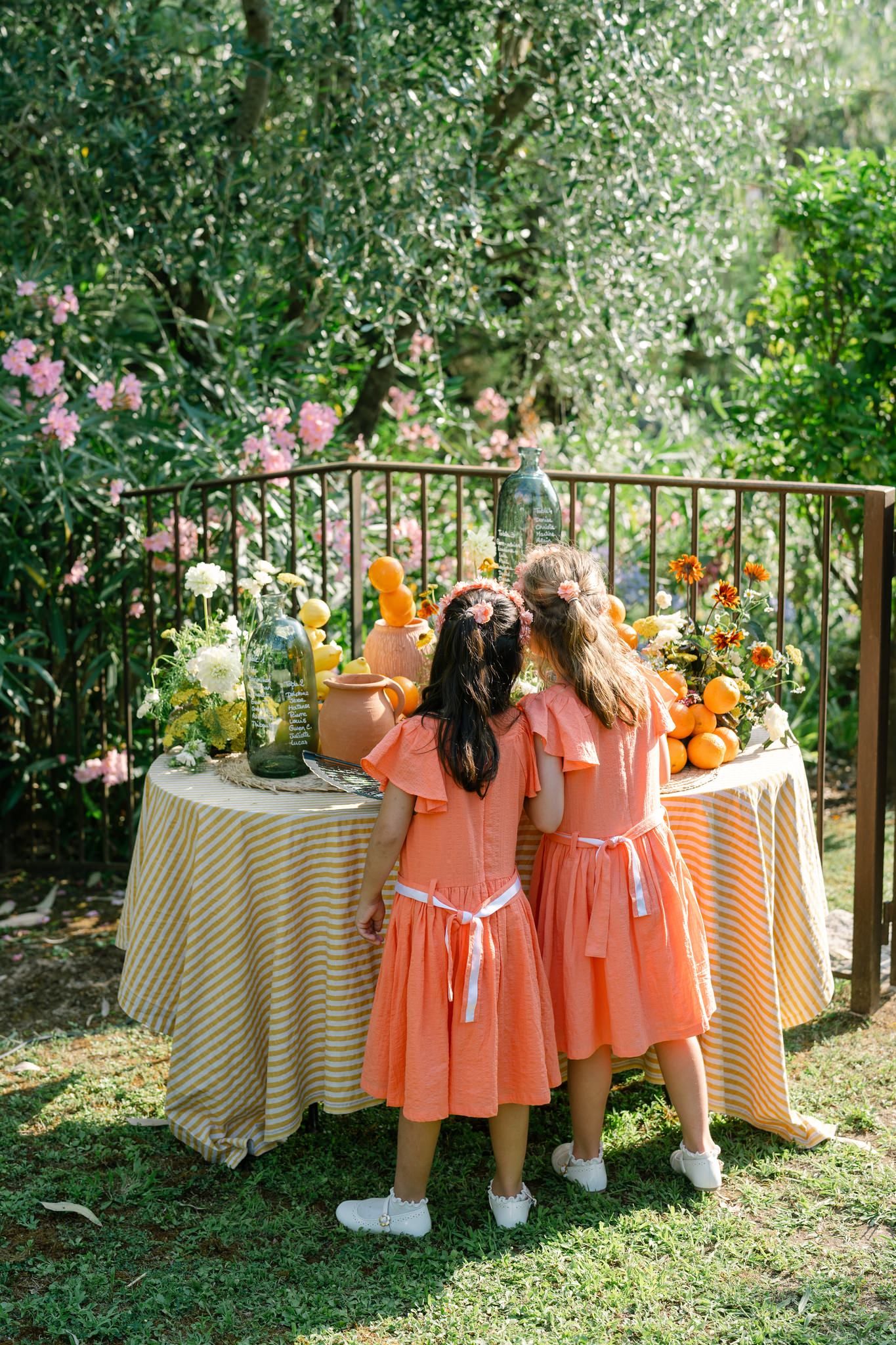 Bridesmaids in coral-colored dresses gather near tables with striped linens in a garden setting.