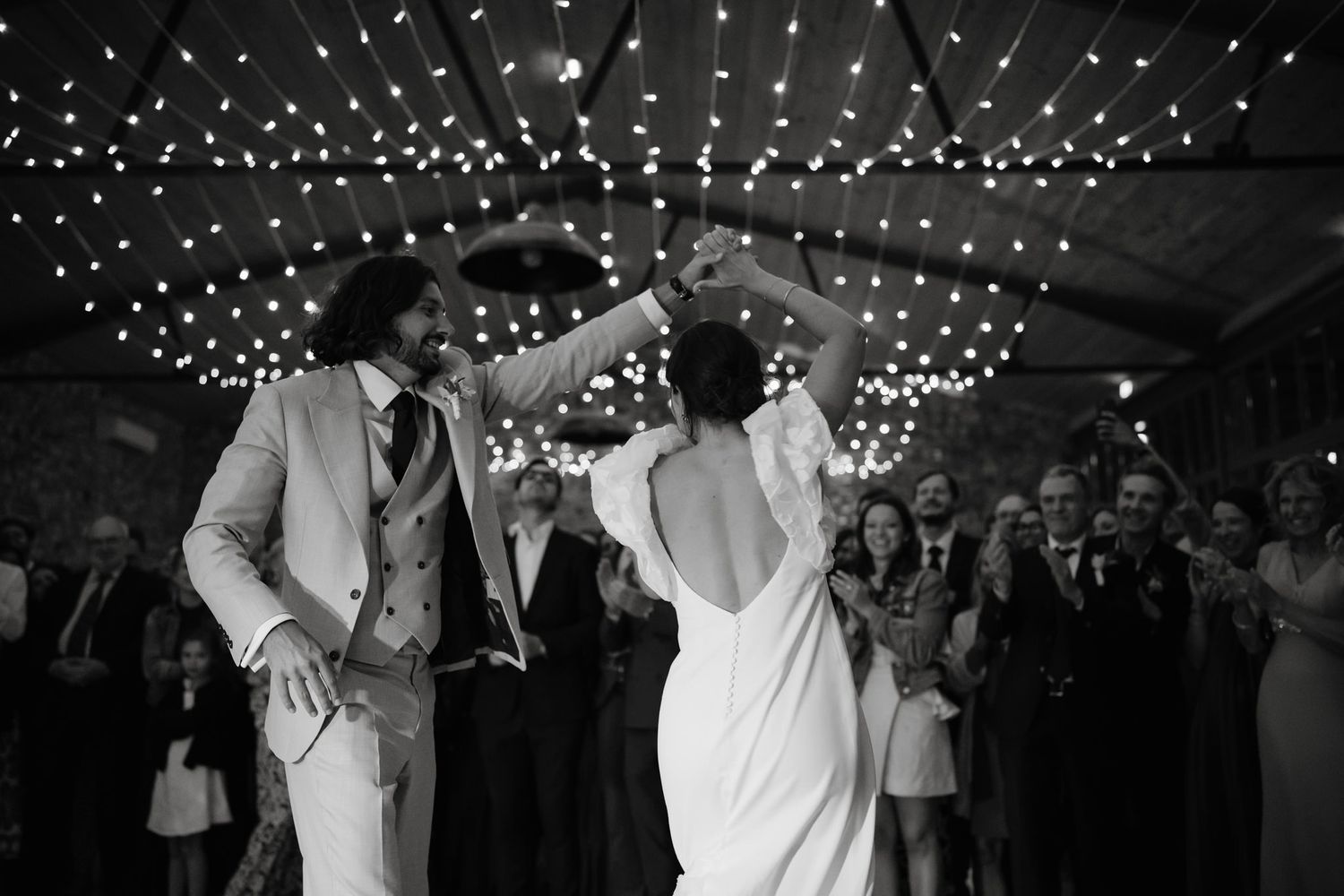 Wedding party dances joyfully under twinkling lights at an evening reception celebration.