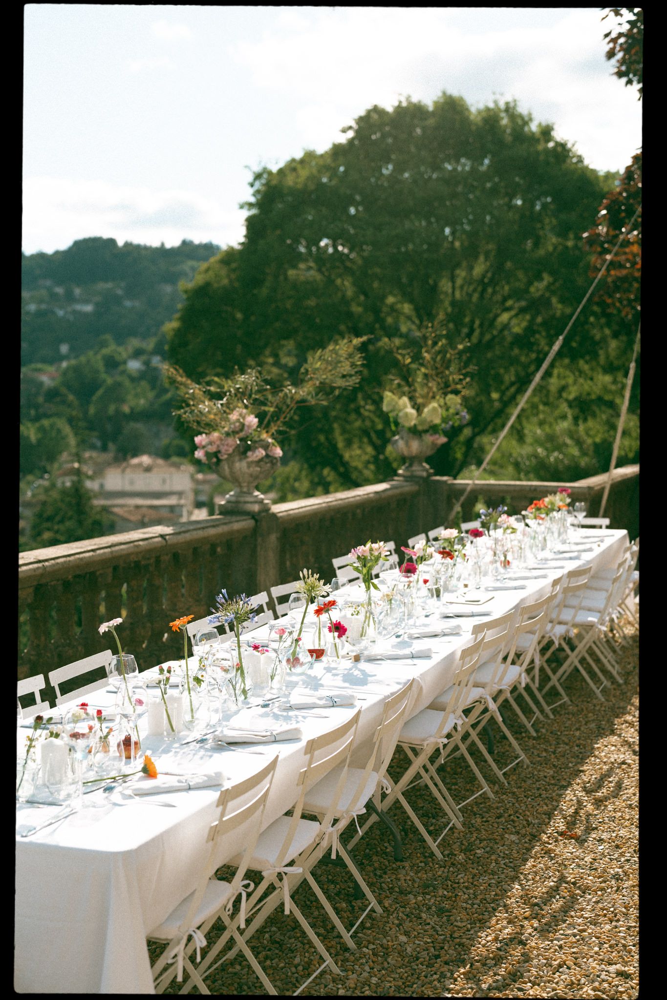 Long white reception tables with place settings arranged outdoors under trees.