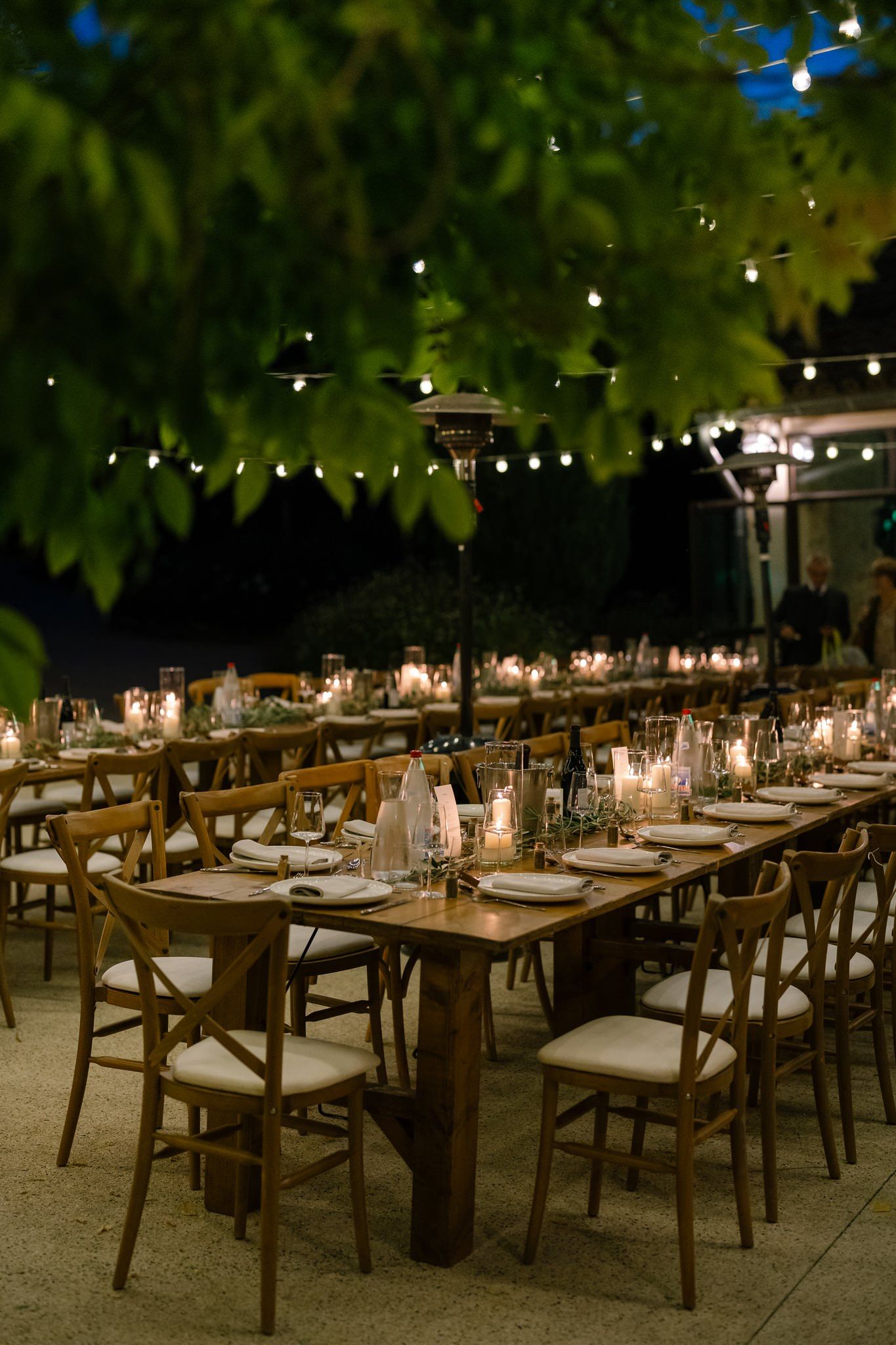 Outdoor wedding reception setup with crossback chairs and string lights overhead at dusk.