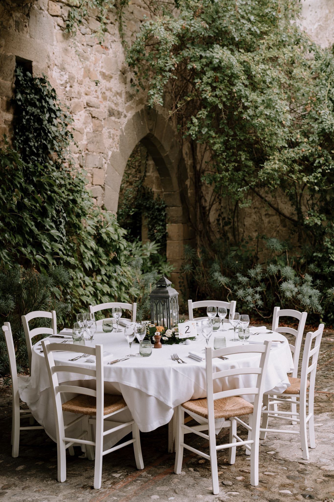 White dining tables and chairs arranged in a romantic stone courtyard with climbing vines.
