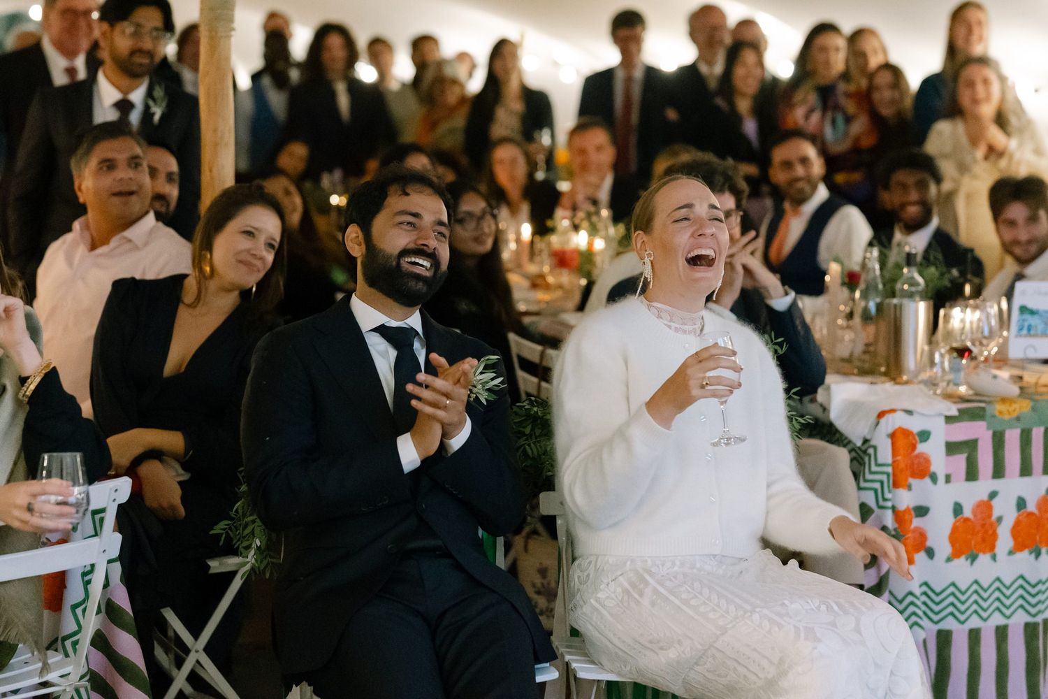 Wedding guests sharing laughter and joy during speeches at an outdoor reception.