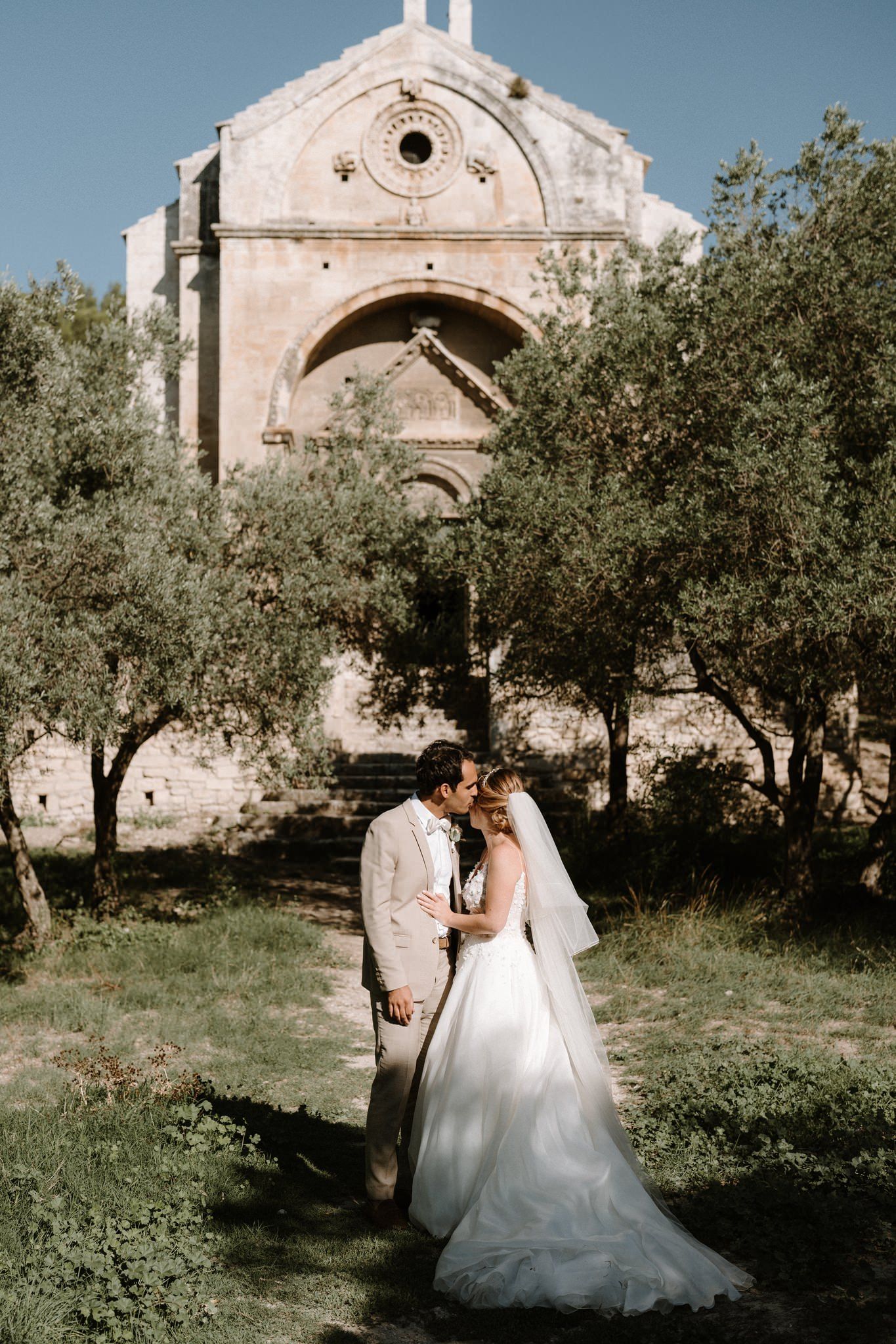 Wedding couple poses for romantic photos in front of historic stone church surrounded by olive trees.