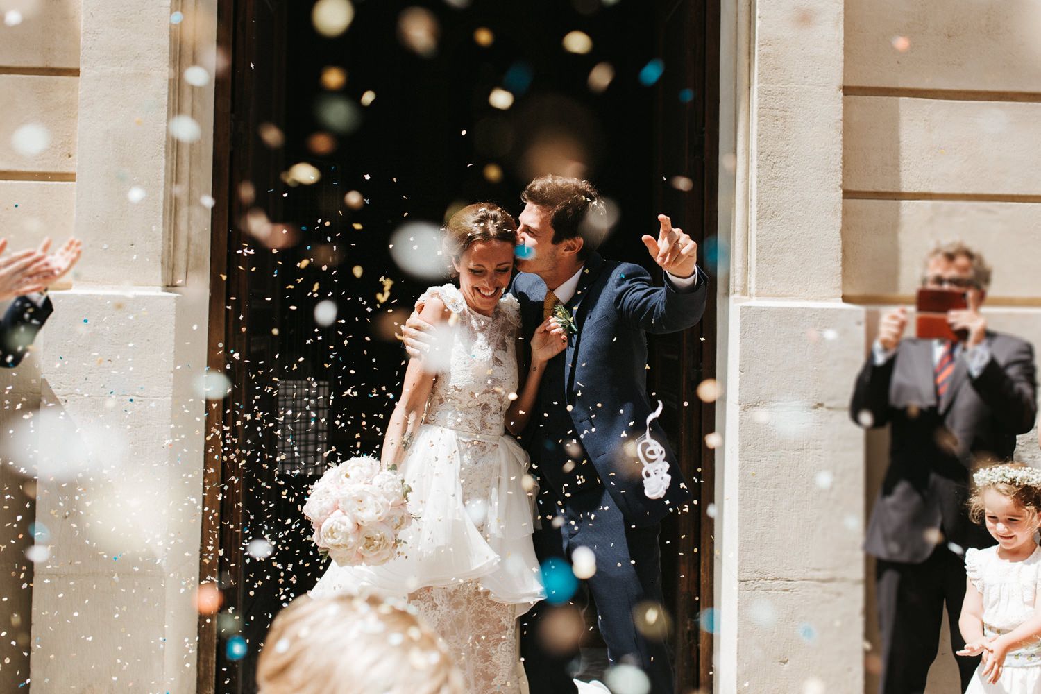 A joyful wedding exit with confetti showering the newlyweds outside a stone building.