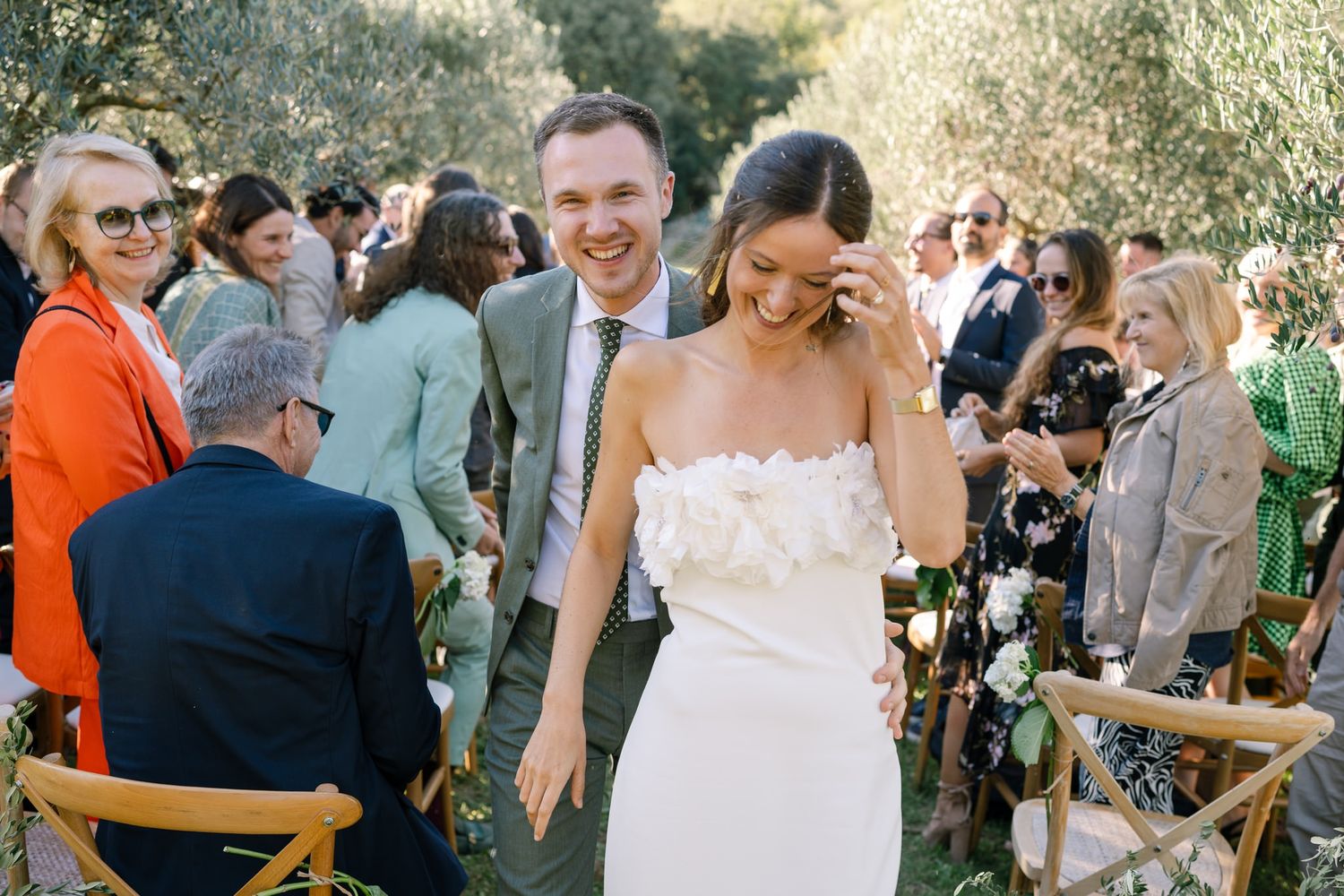Wedding celebration with guests seated at long tables among olive trees.