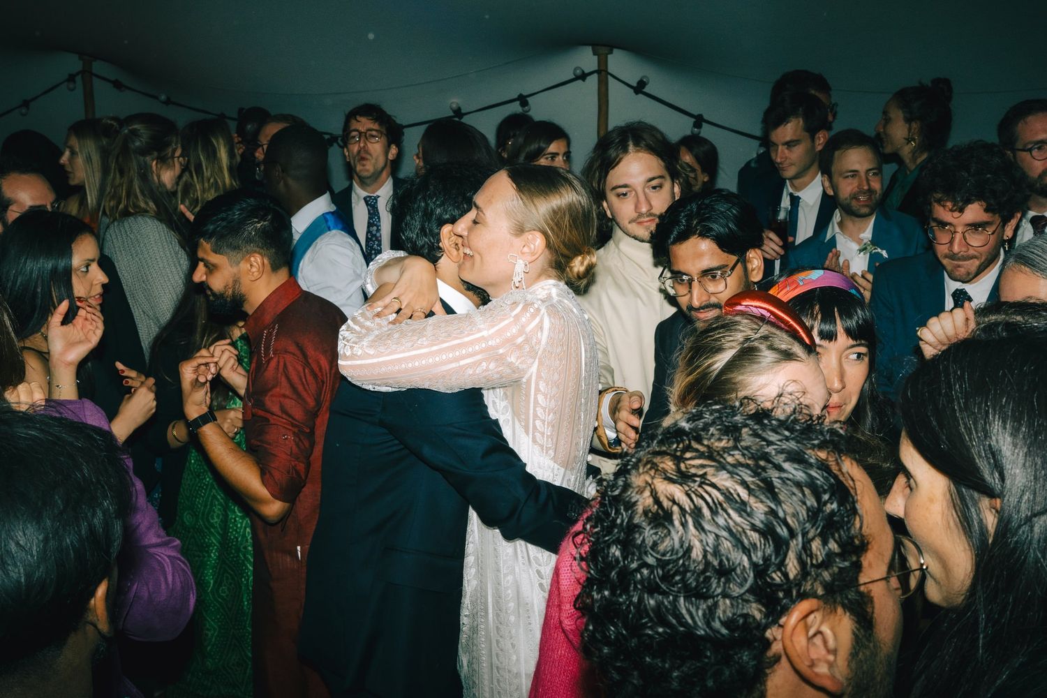 Wedding guests dancing and embracing on a dimly lit dance floor during celebration.