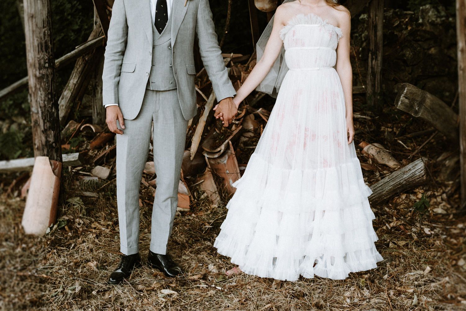 A wedding couple poses together in a wooded outdoor setting wearing a white gown and light gray suit.