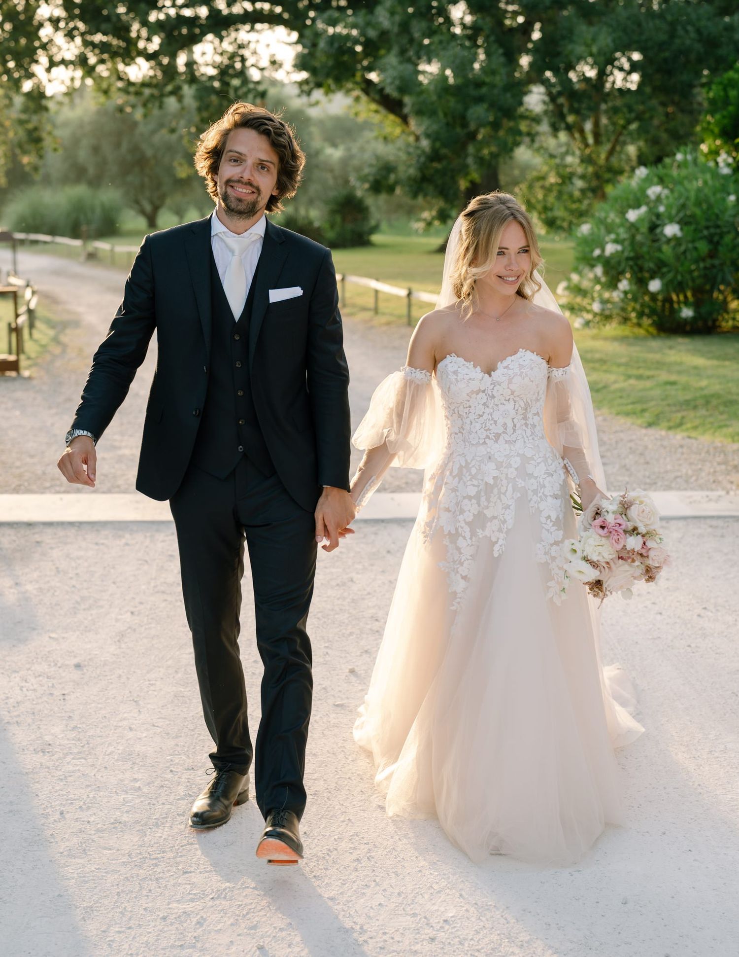 Wedding couple strolls along a white gravel path surrounded by lush greenery.