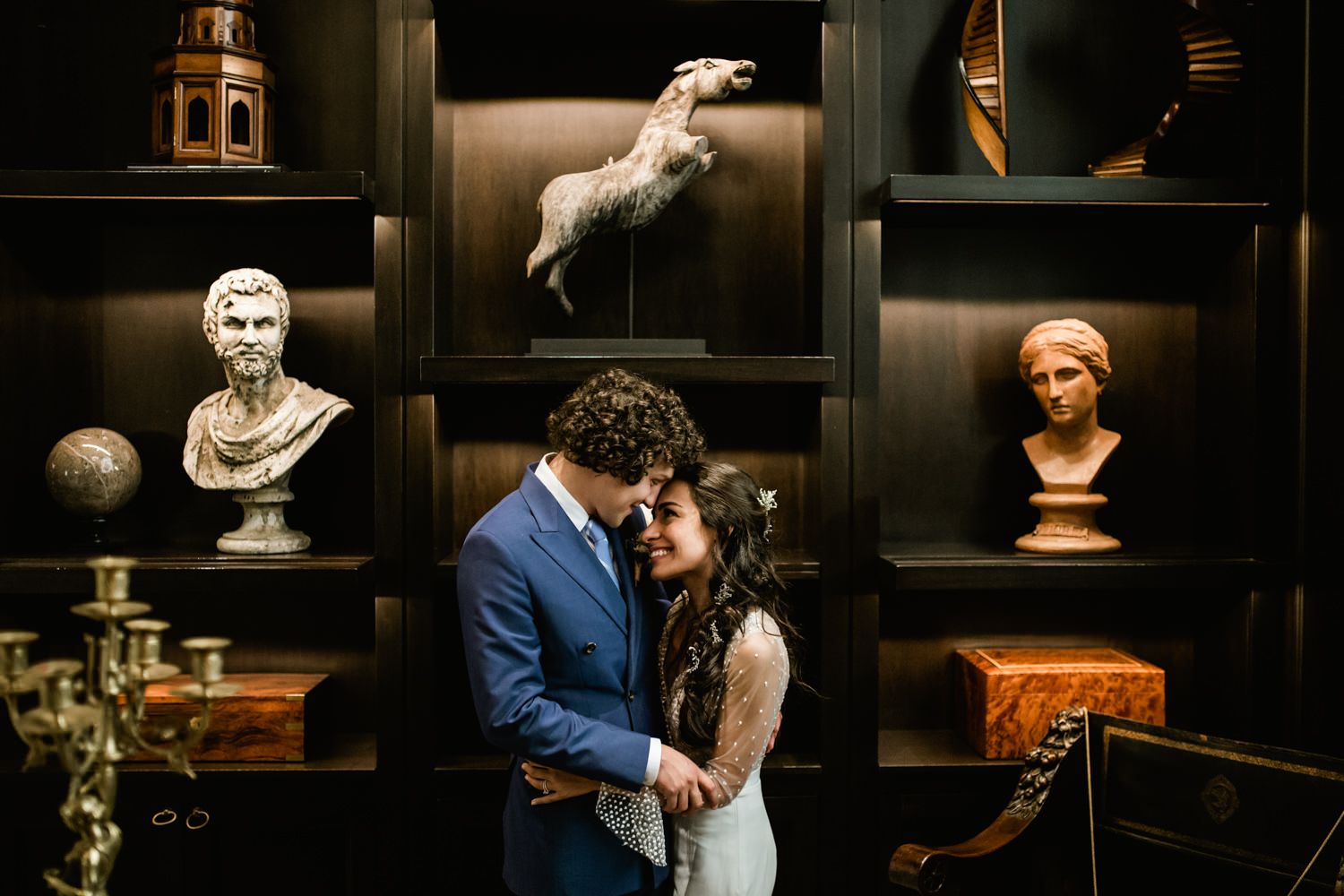 Couple embracing in an elegant room decorated with classical busts and vintage artifacts.