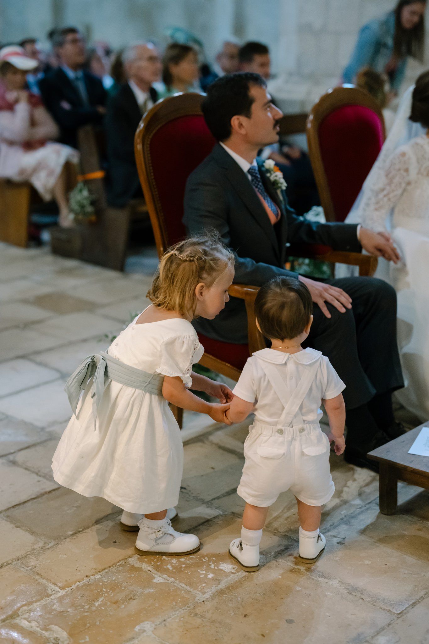 Two young children in white formal wear play near red velvet chairs during ceremony.
