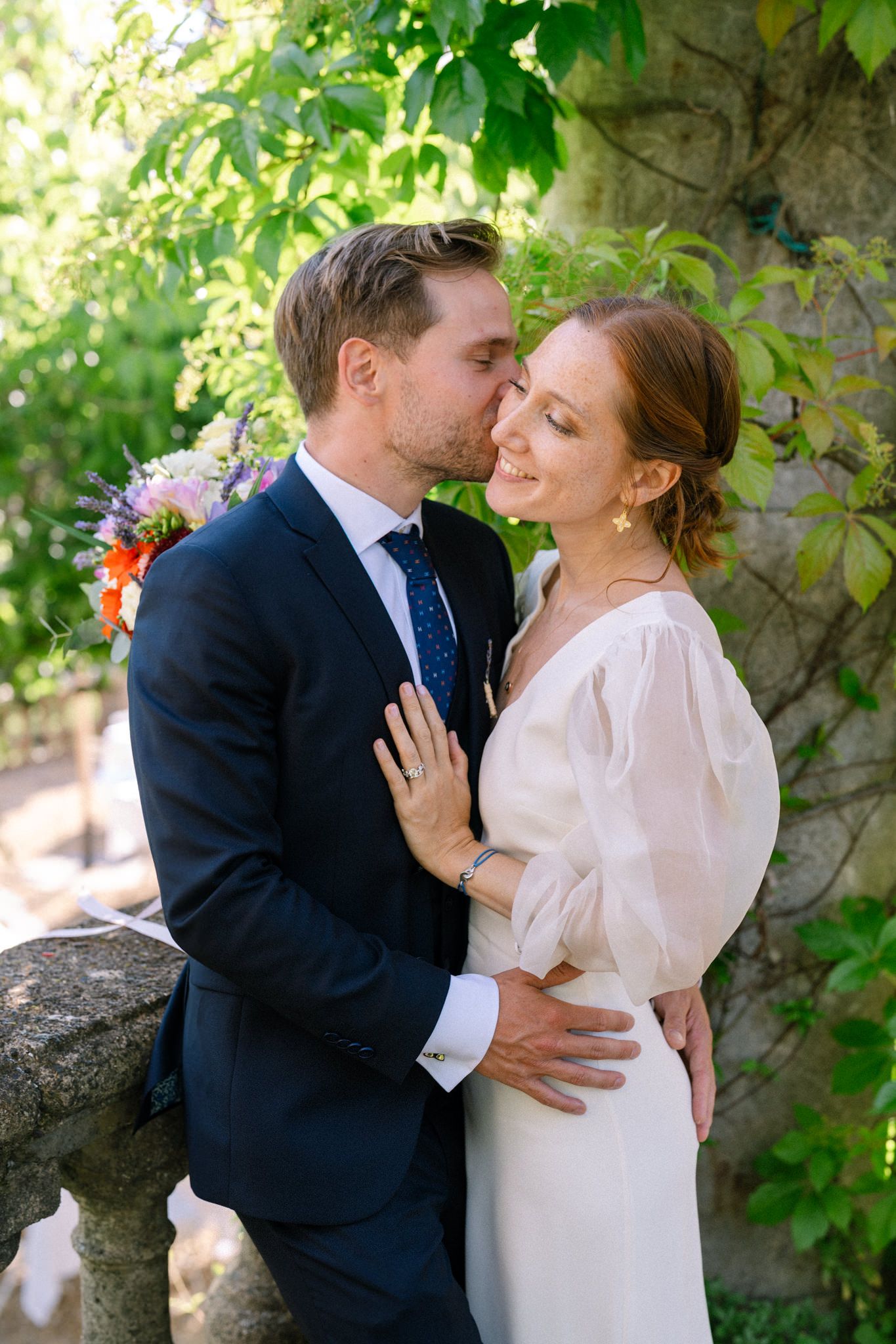 A tender moment between newlyweds in formal attire against a garden wall backdrop.
