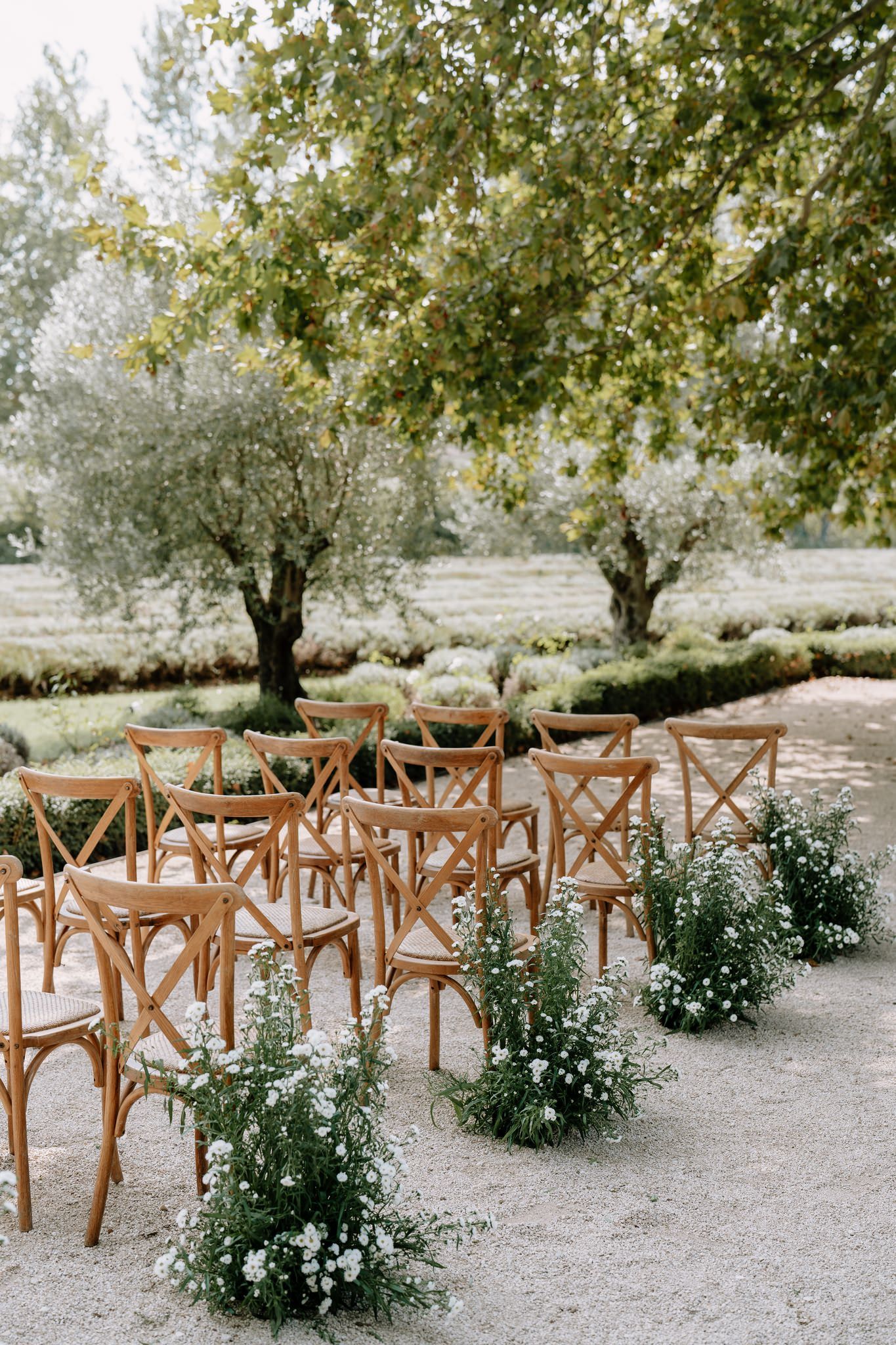 Rustic outdoor wedding ceremony setup with wooden cross-back chairs and white floral arrangements.