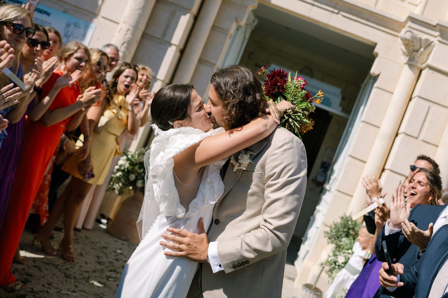 Wedding couple shares an intimate moment outside a grand building as guests look on.