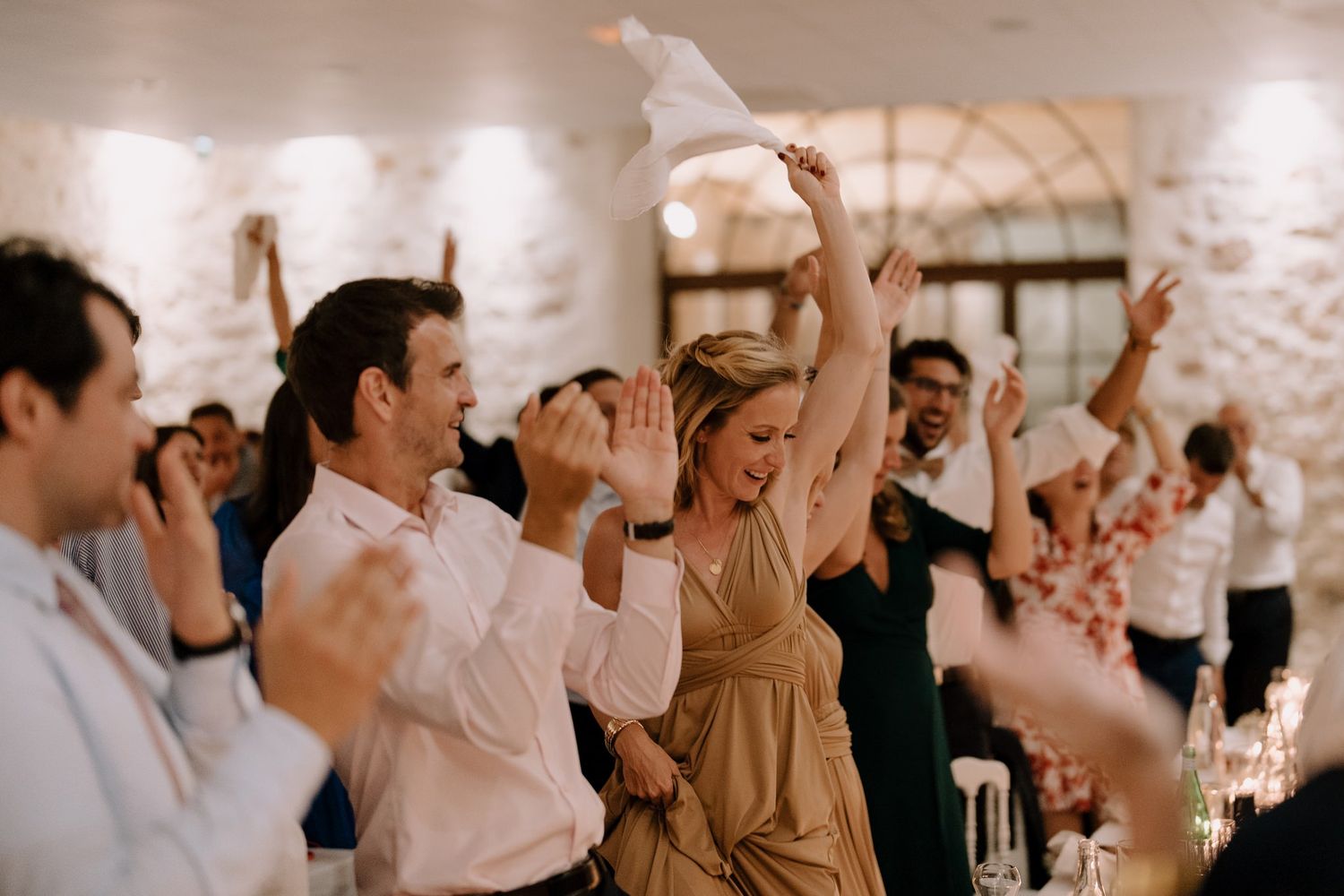 Wedding reception crowd cheering and celebrating with raised hands in a stone-walled venue.