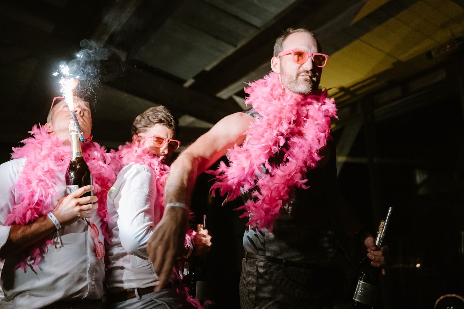 Wedding party members dancing with pink feather boas under dramatic lighting.