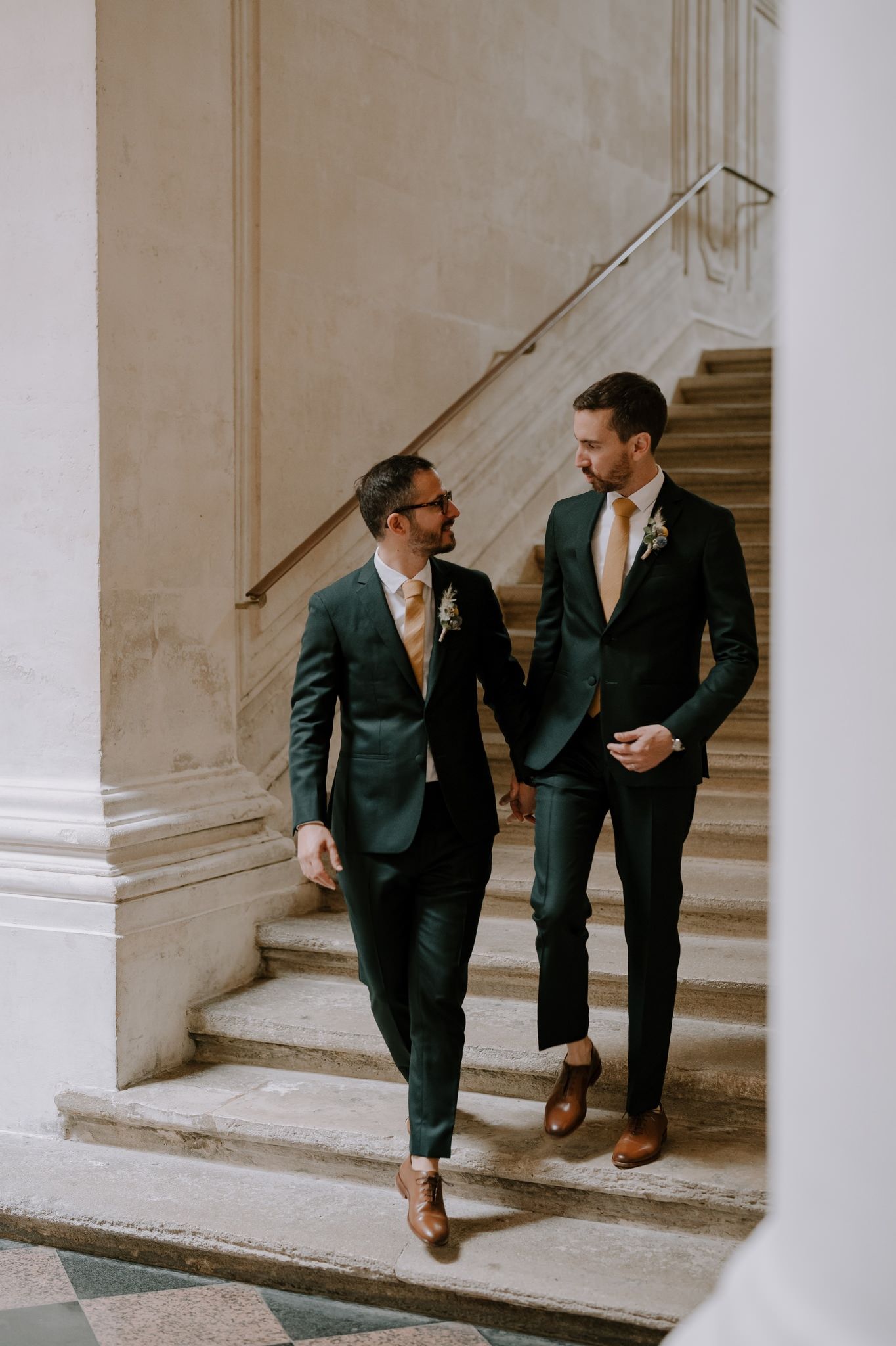 Three men in dark suits walk up stone steps in an elegant interior.