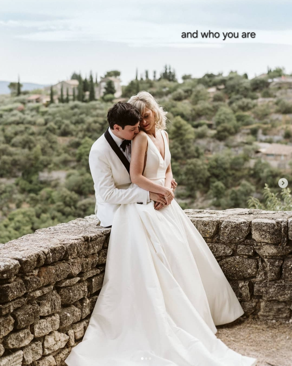 A couple in wedding attire embraces while standing on a stone wall overlooking a scenic mountain landscape.