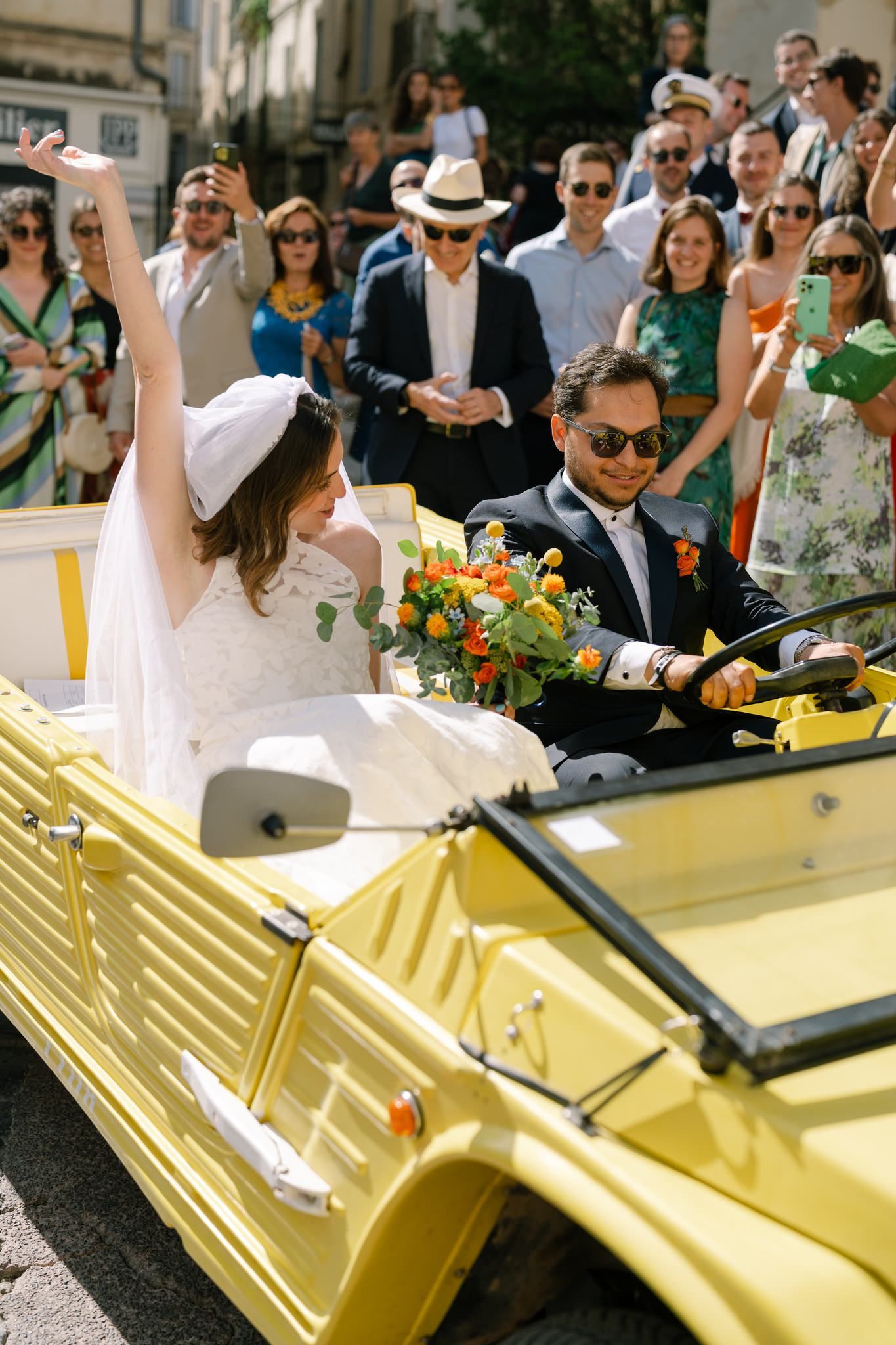 Newlyweds riding in a vintage yellow convertible car surrounded by wedding guests.