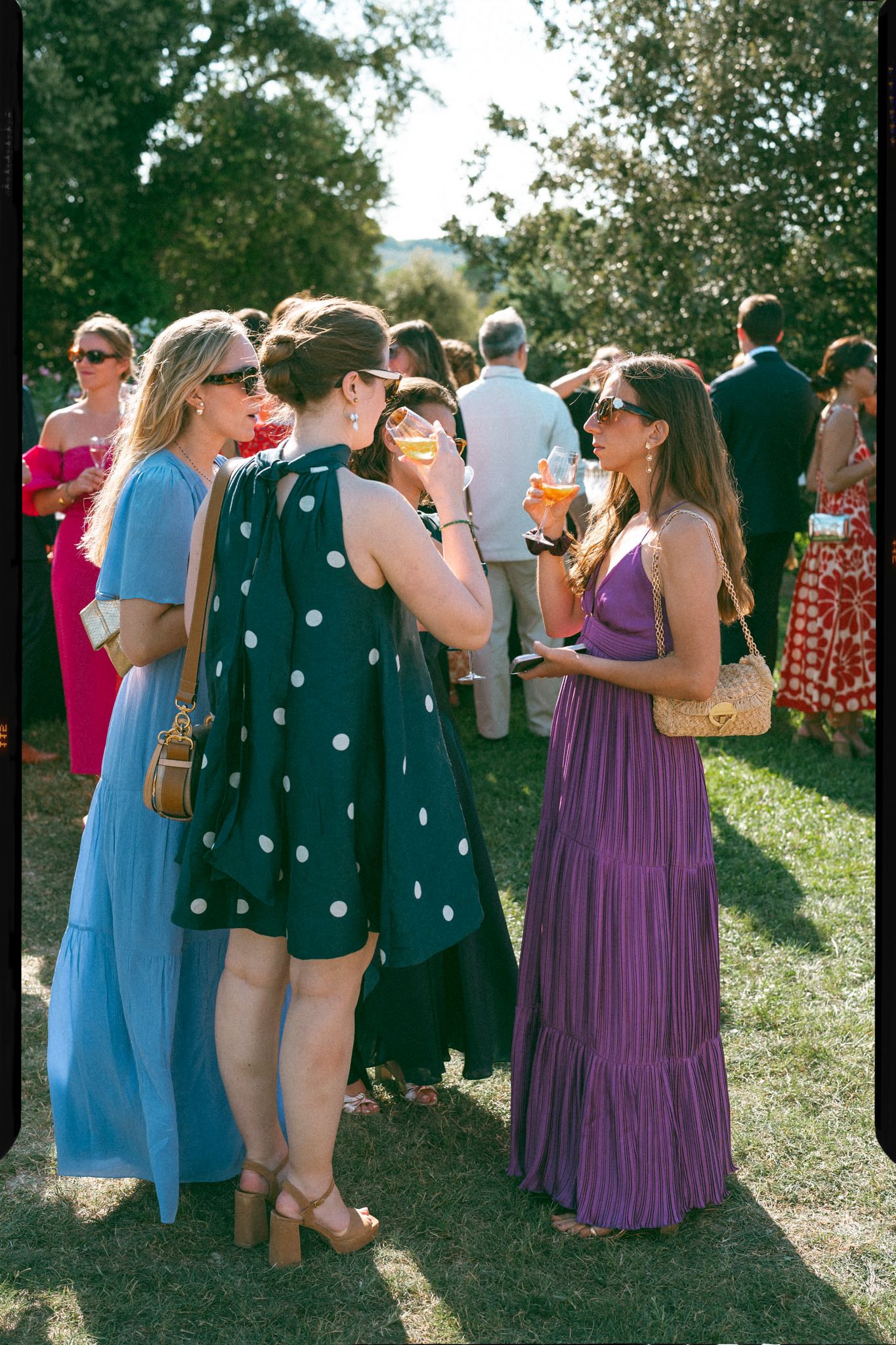 Wedding guests in blue and purple dresses mingle outdoors during a summer celebration.