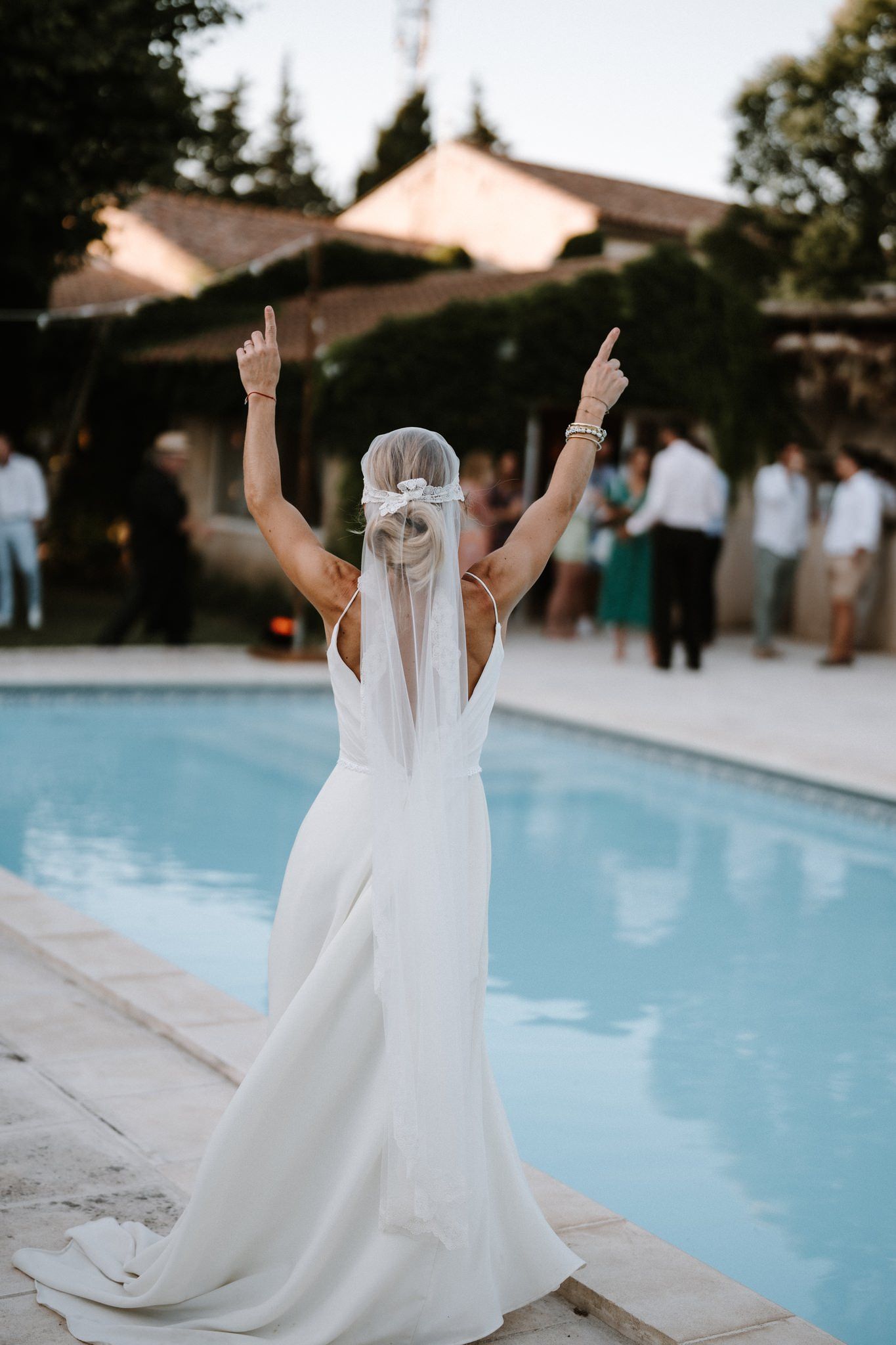 Bride in elegant white dress raises her arms joyfully by a swimming pool during outdoor wedding reception.