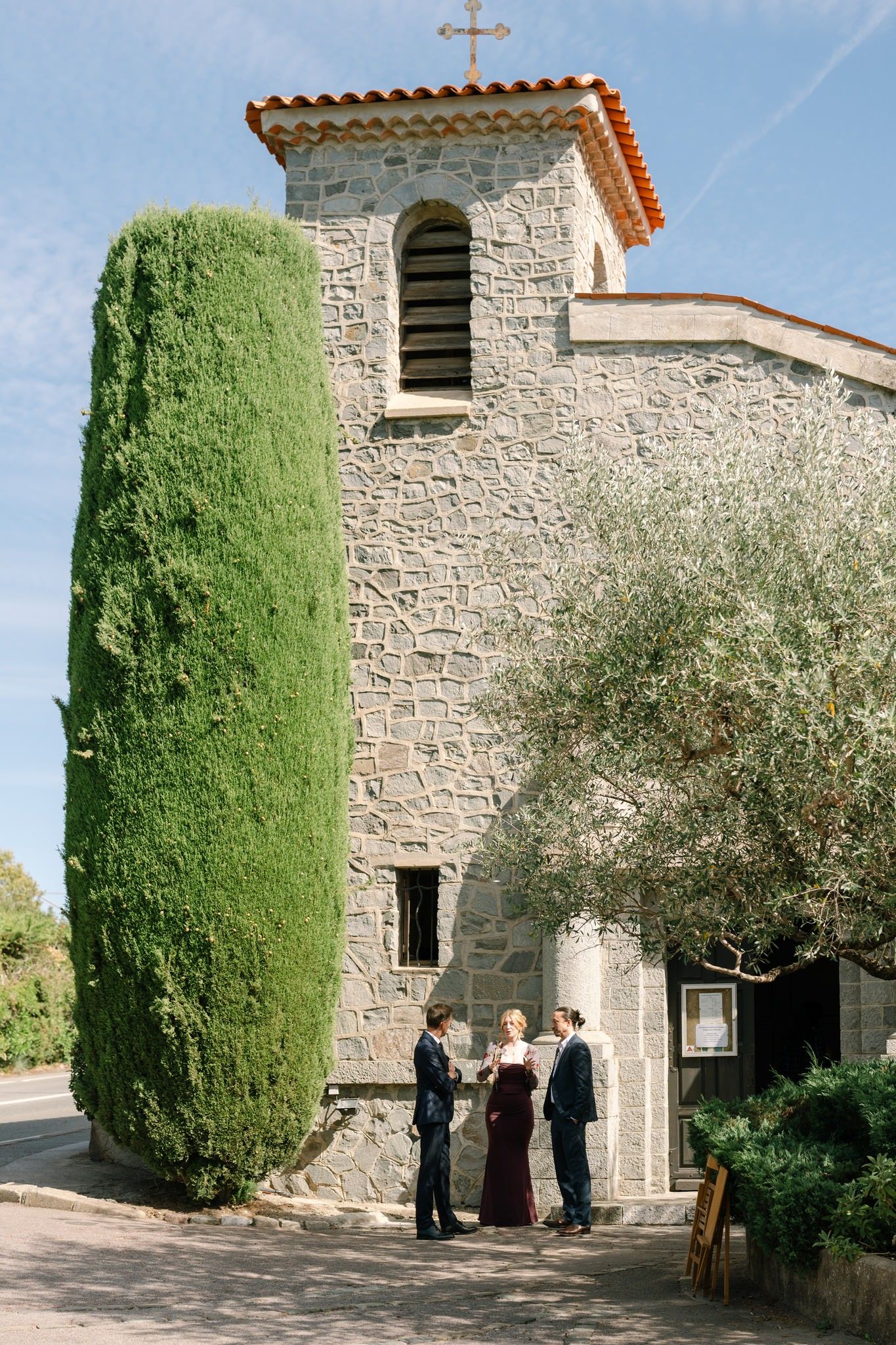 Stone chapel exterior with tall cypress trees and Mediterranean architecture.