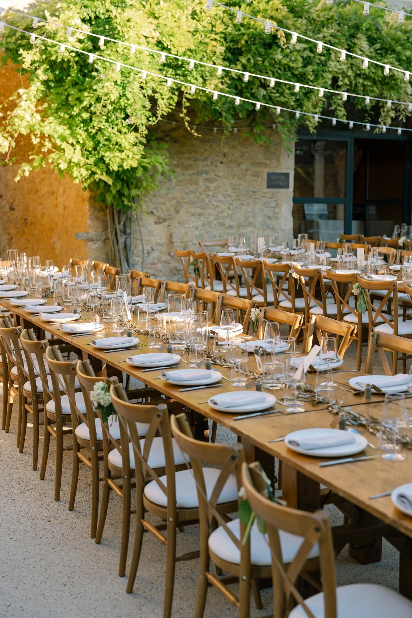 Long wooden tables set for outdoor wedding reception under string lights and trees.