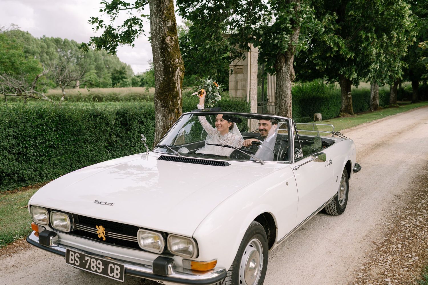 Vintage white convertible drives along tree-lined path at wedding venue.