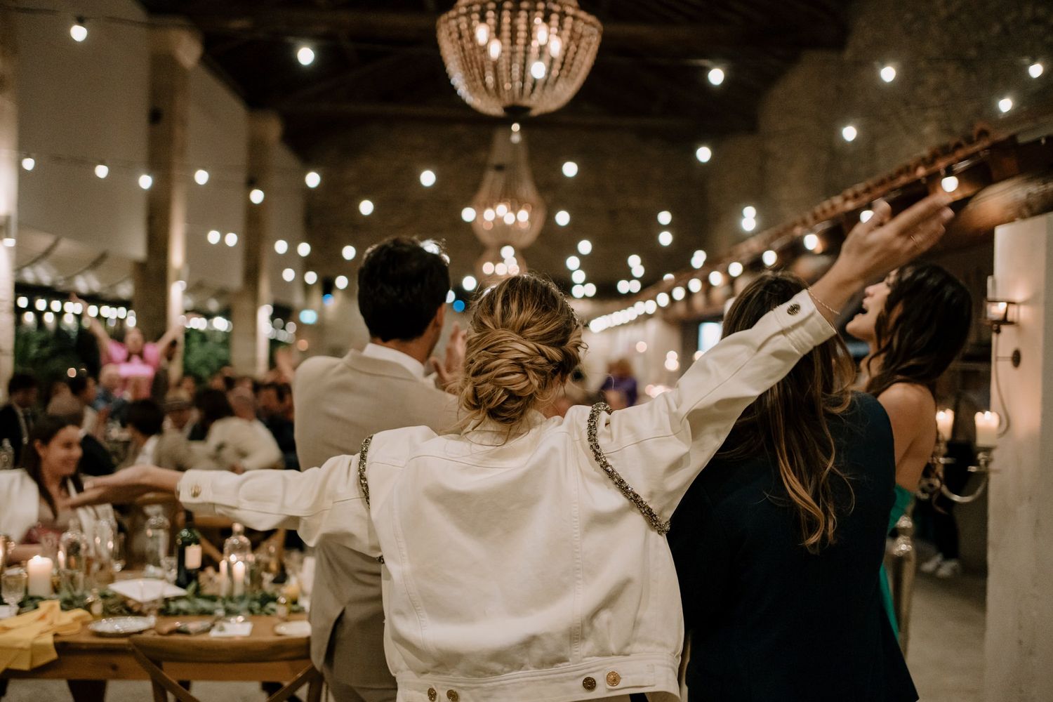 Wedding celebration with string lights illuminating guests dancing with raised arms.