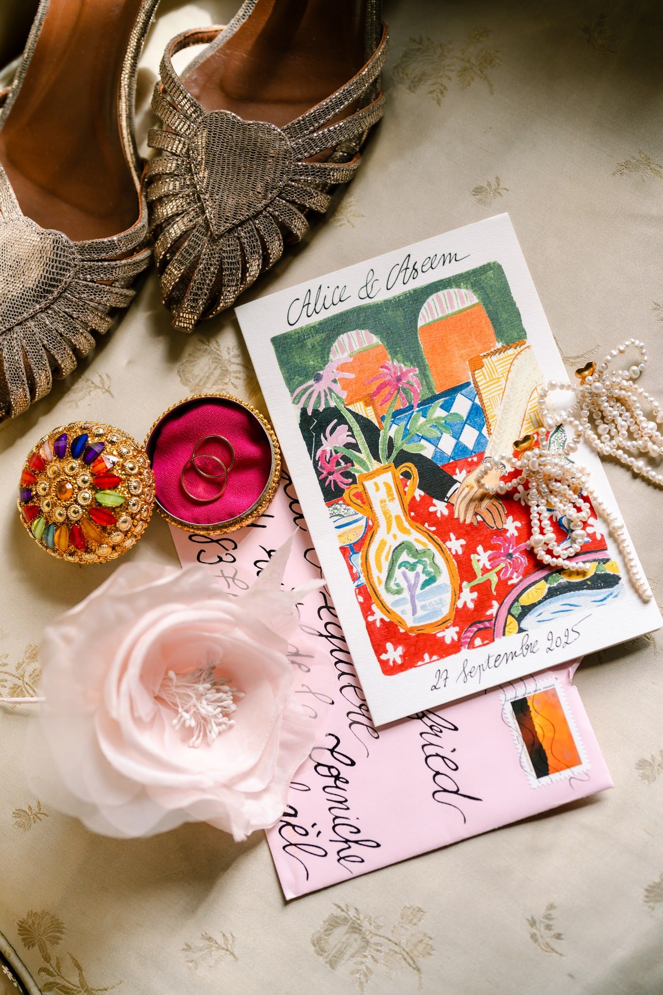 Wedding details including vintage shoes, colorful invitation card, and delicate floral accessories arranged on light surface.