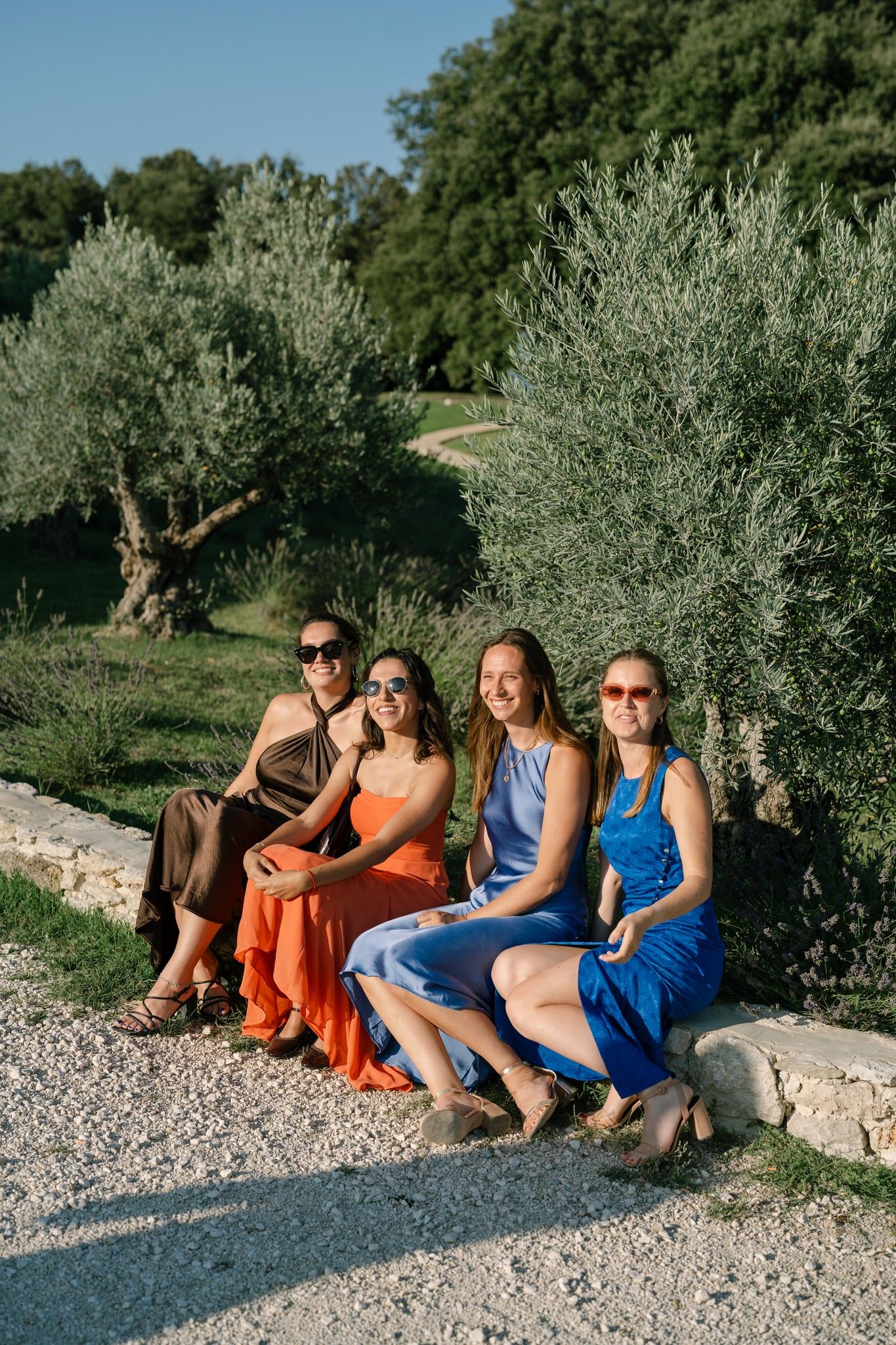Group relaxing on stone wall among olive trees wearing bright summer dresses.