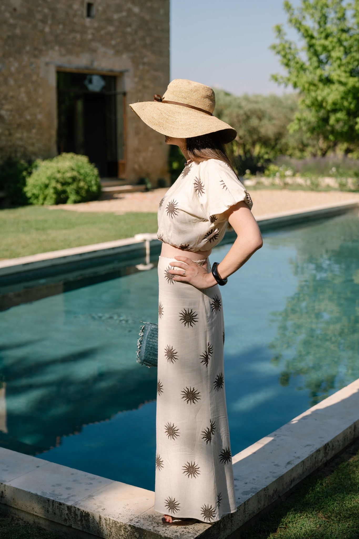 Figure in patterned dress and sun hat poses by modern swimming pool.