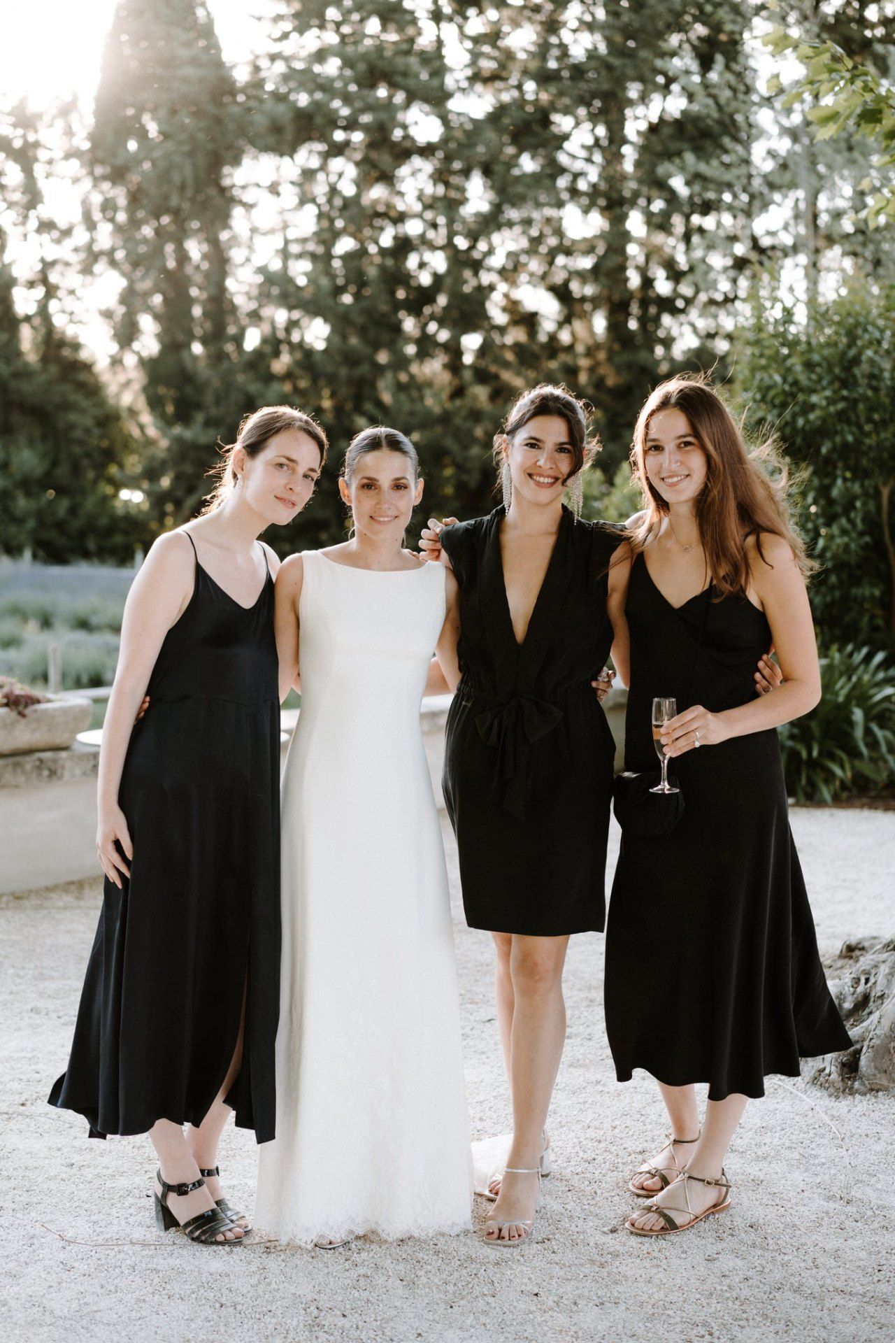 Bride in white gown poses with three bridesmaids in black dresses outdoors at a wedding celebration.