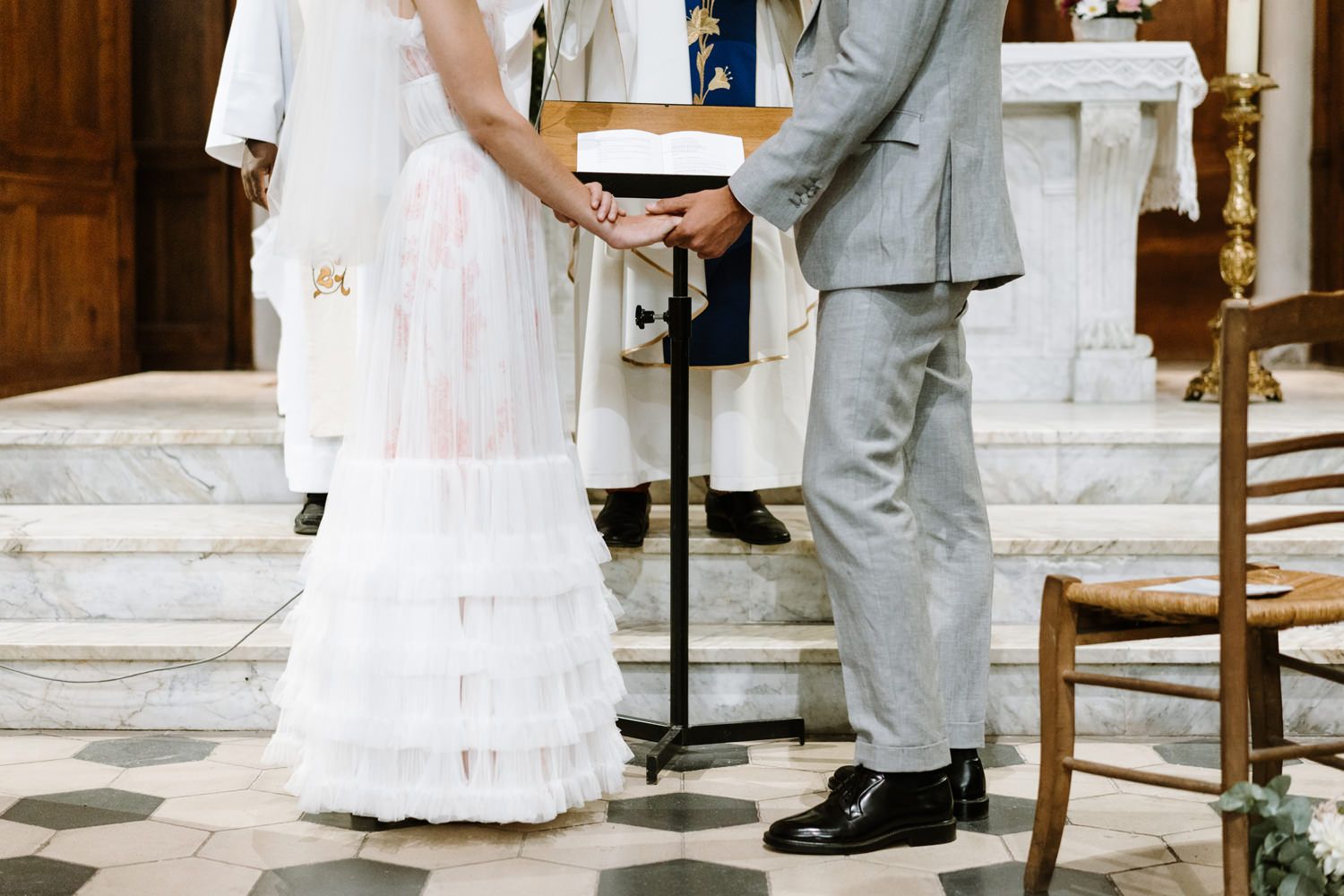 A close-up view of a bride in white dress and groom in gray suit holding hands during a church ceremony.