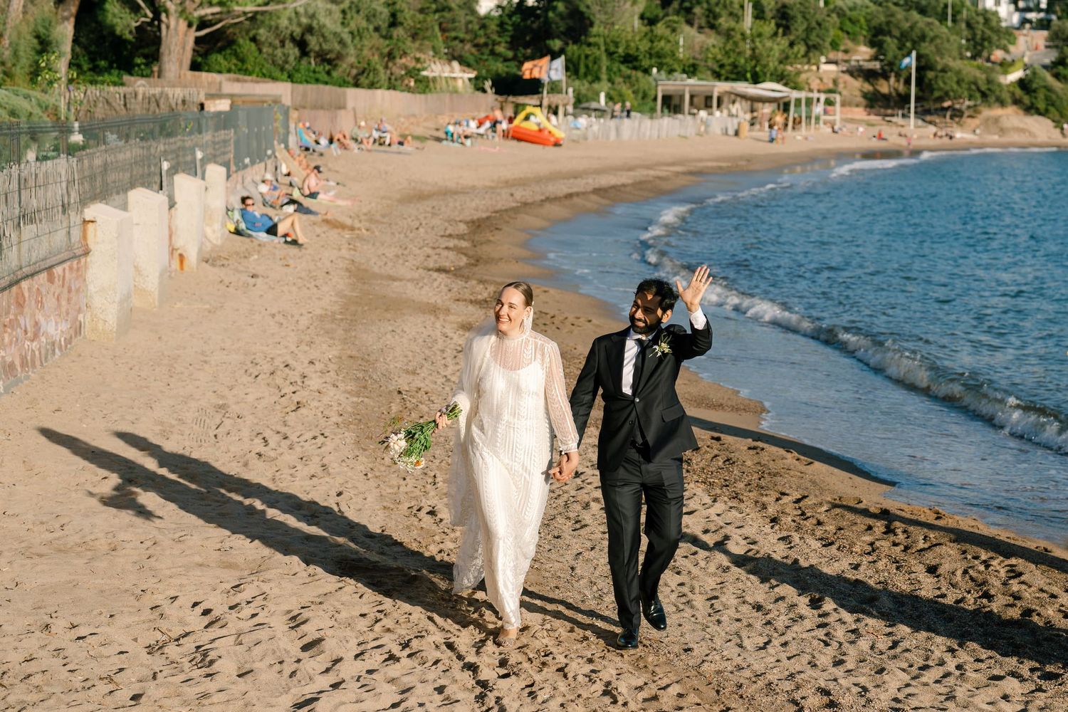 Wedding couple strolling on a sandy Mediterranean beach with cliffs in the distance.