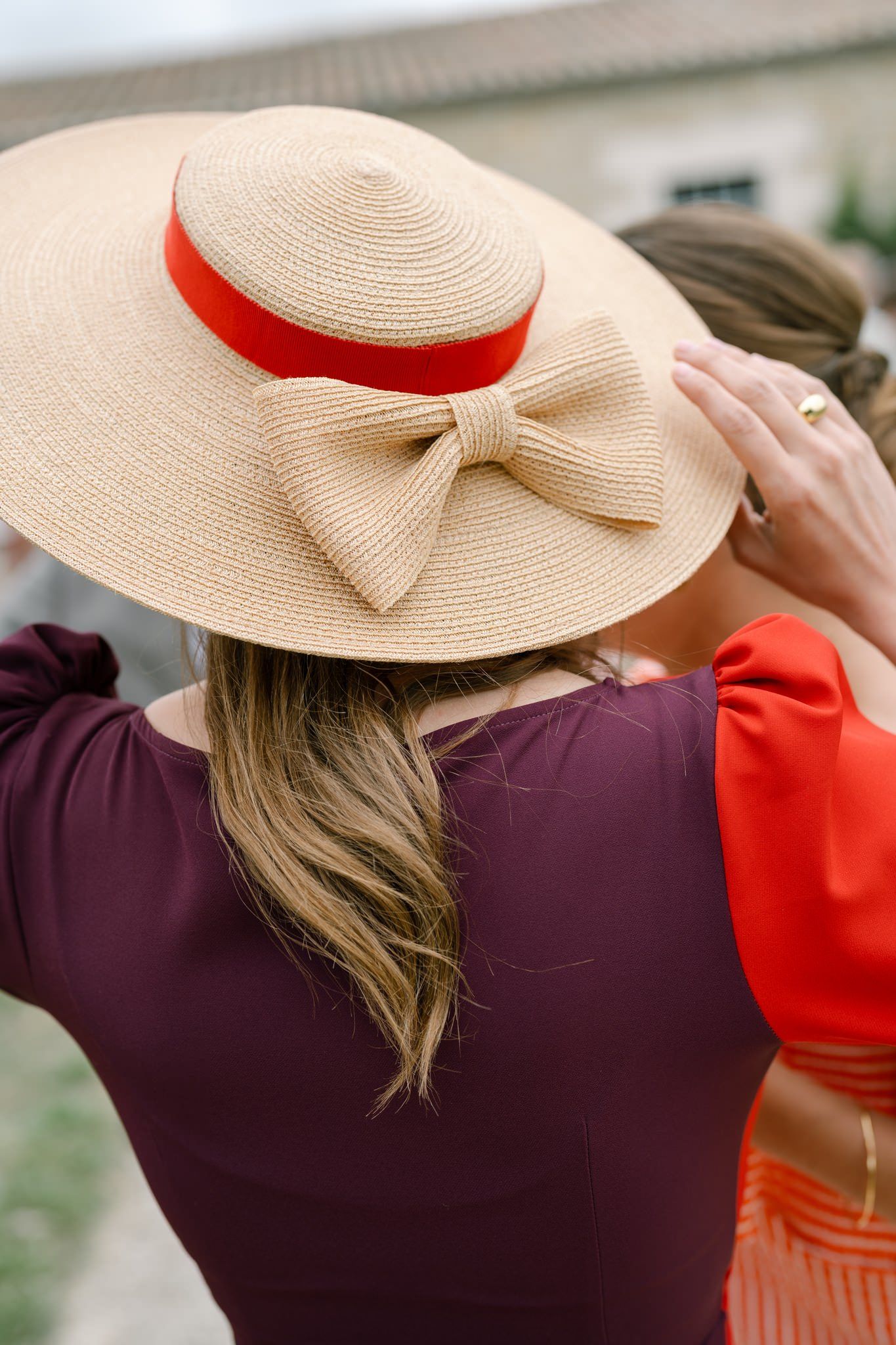 Woman in burgundy dress and straw sun hat with red ribbon shown from behind in outdoor setting.