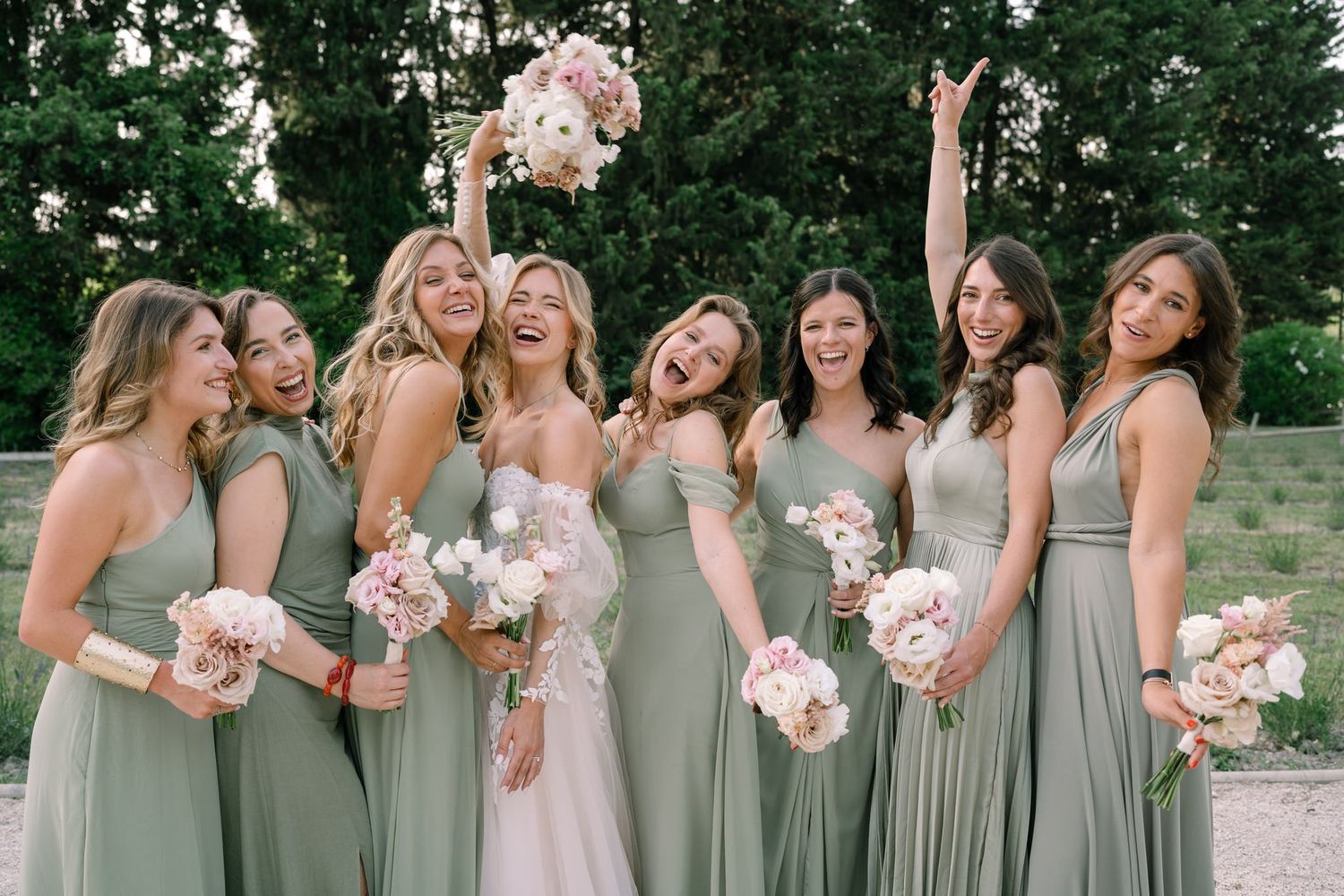 Bridesmaids in sage green dresses stand in a row holding pink bouquets at an outdoor wedding.