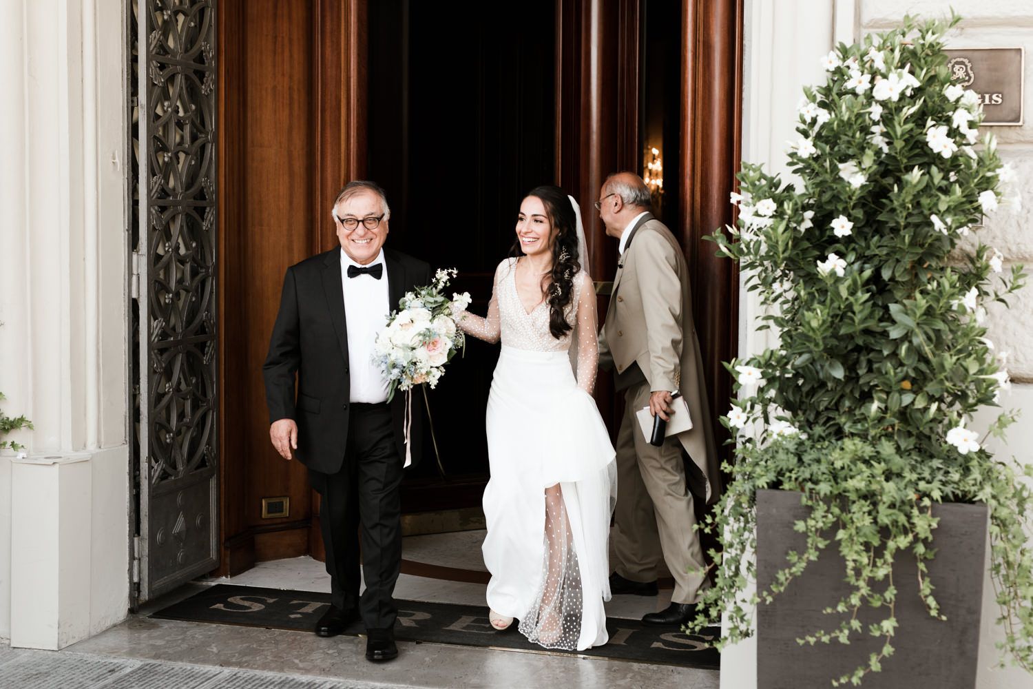 Wedding party emerges from elegant doorway with floral arrangements.