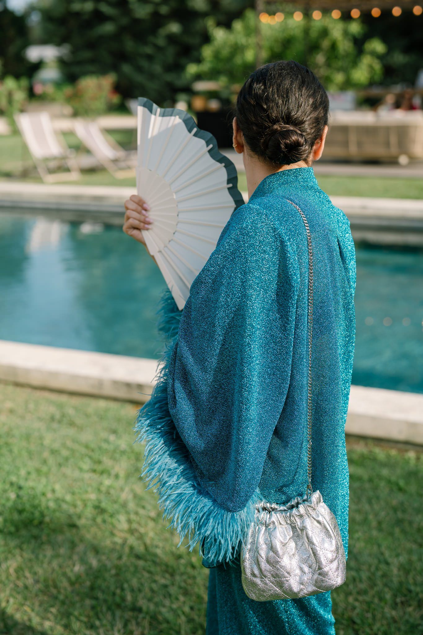 Person wearing a sparkly turquoise shawl holding a white fan by a swimming pool.
