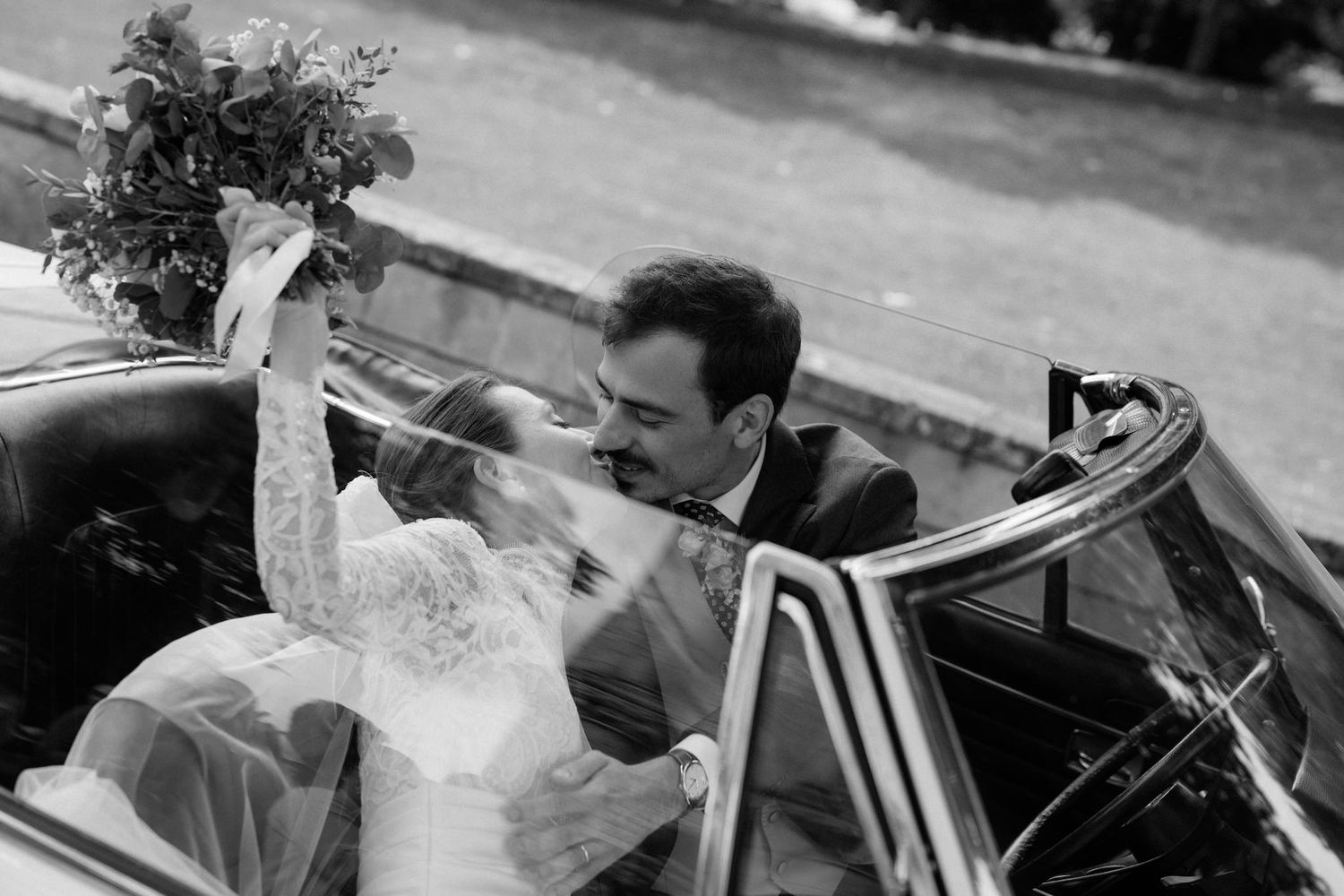 Black and white photo of couple waving from classic car after ceremony.