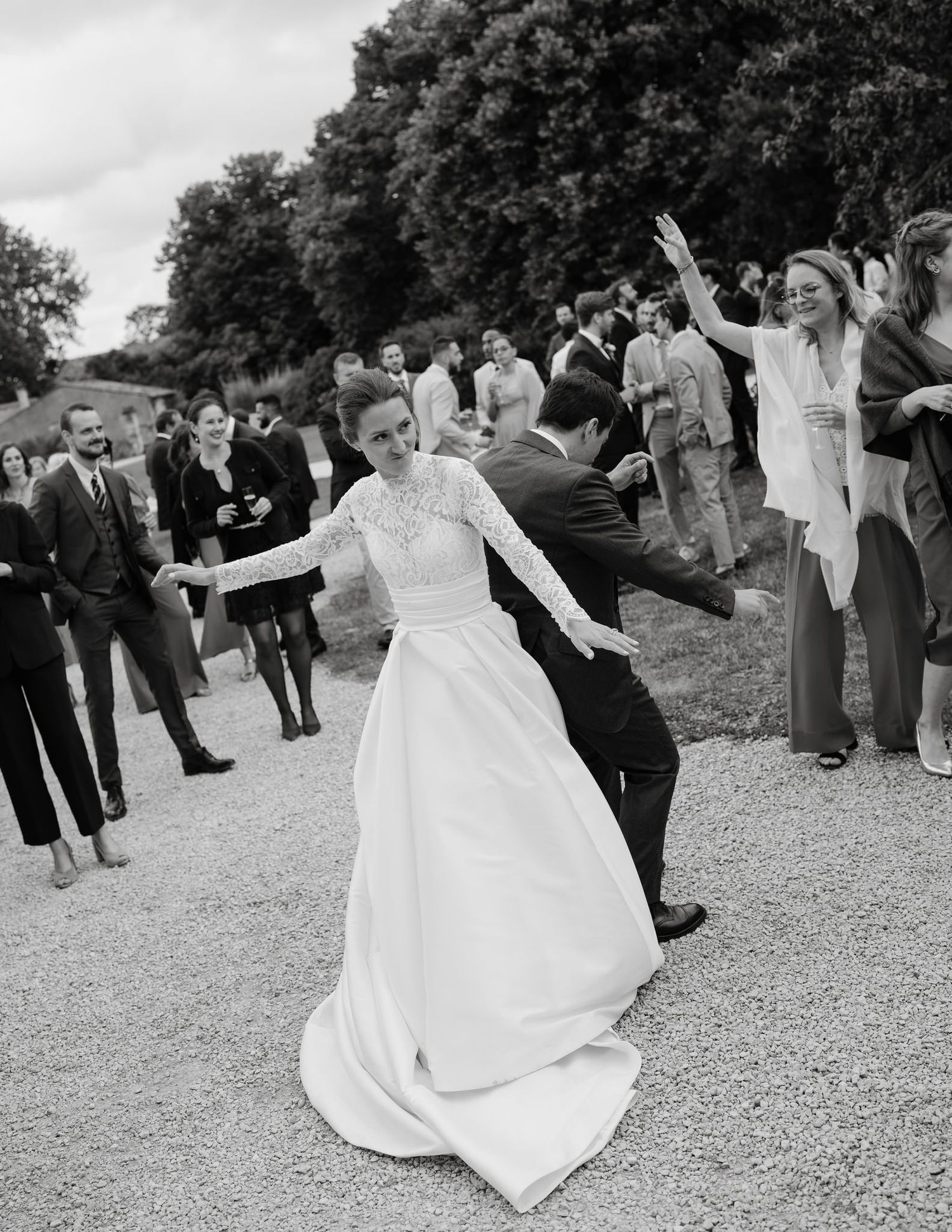 A bride in a flowing white gown spins and dances outdoors on a gravel path.
