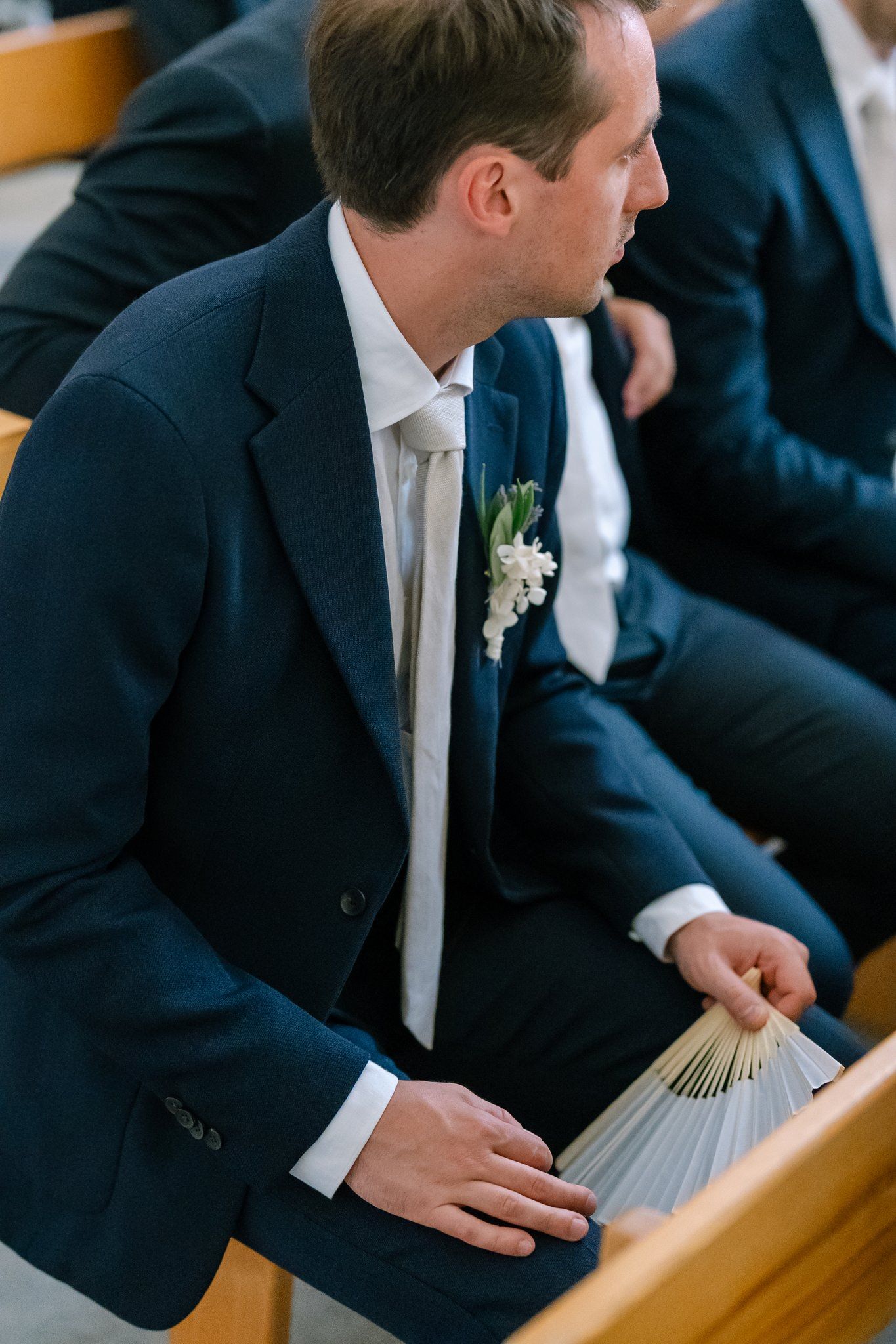 A guest in a navy suit holds a paper fan while seated in a church pew.