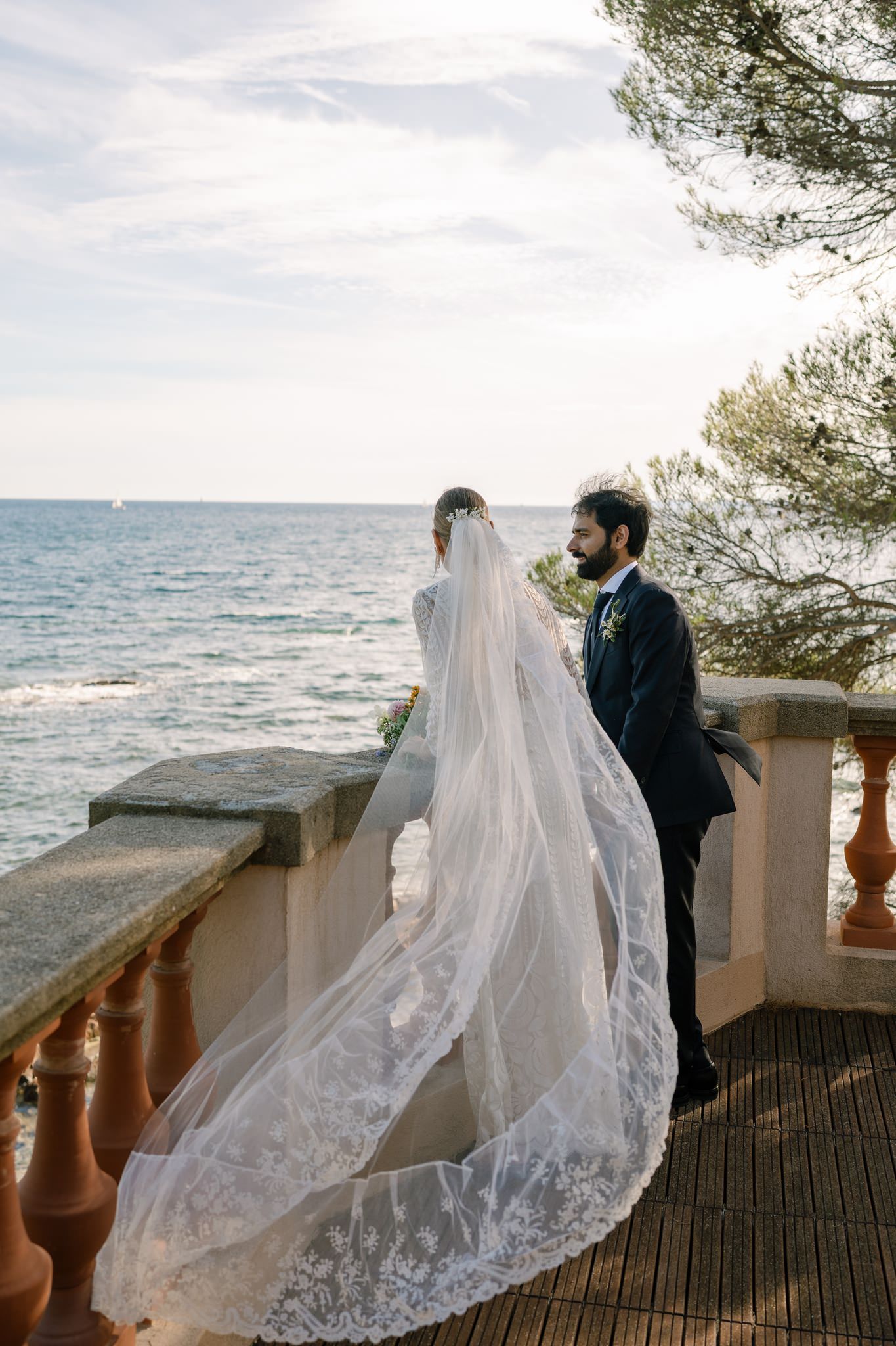A flowing white wedding veil catches the breeze against a coastal backdrop with ornate stone balustrades.