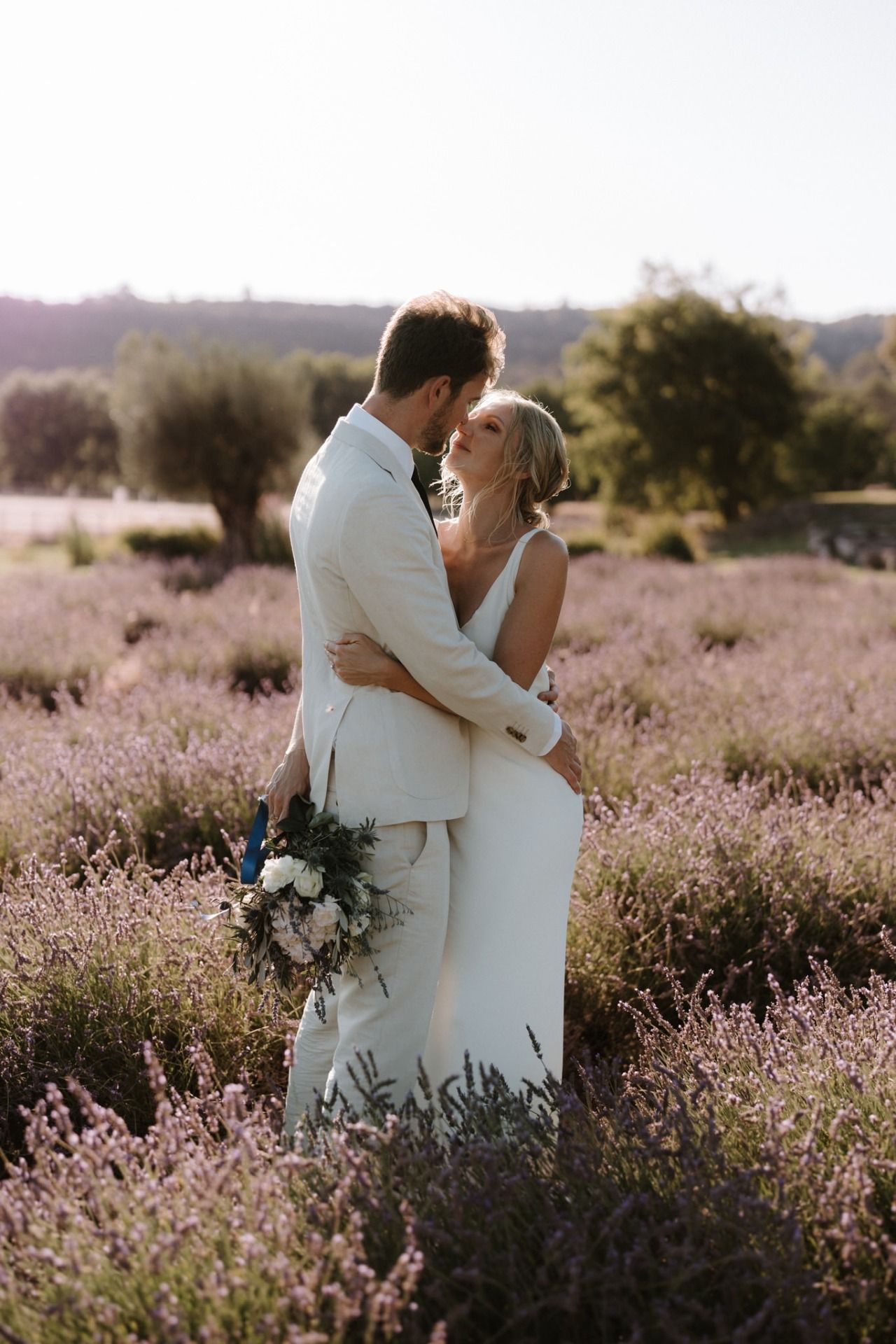 Bride and groom embracing in a lavender field, sharing a romantic moment in matching white wedding attire.