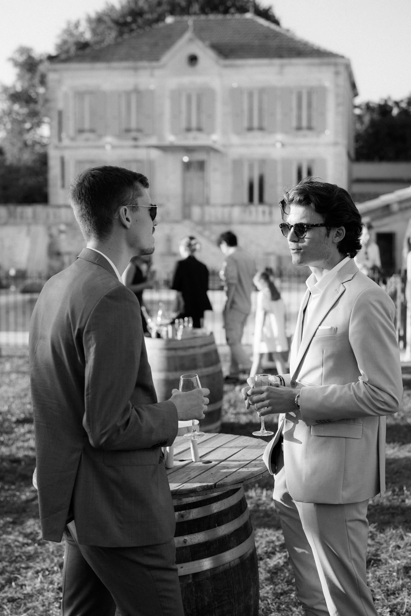 Two guests in sunglasses having a conversation over wine at an elegant outdoor event with a French chateau in background.