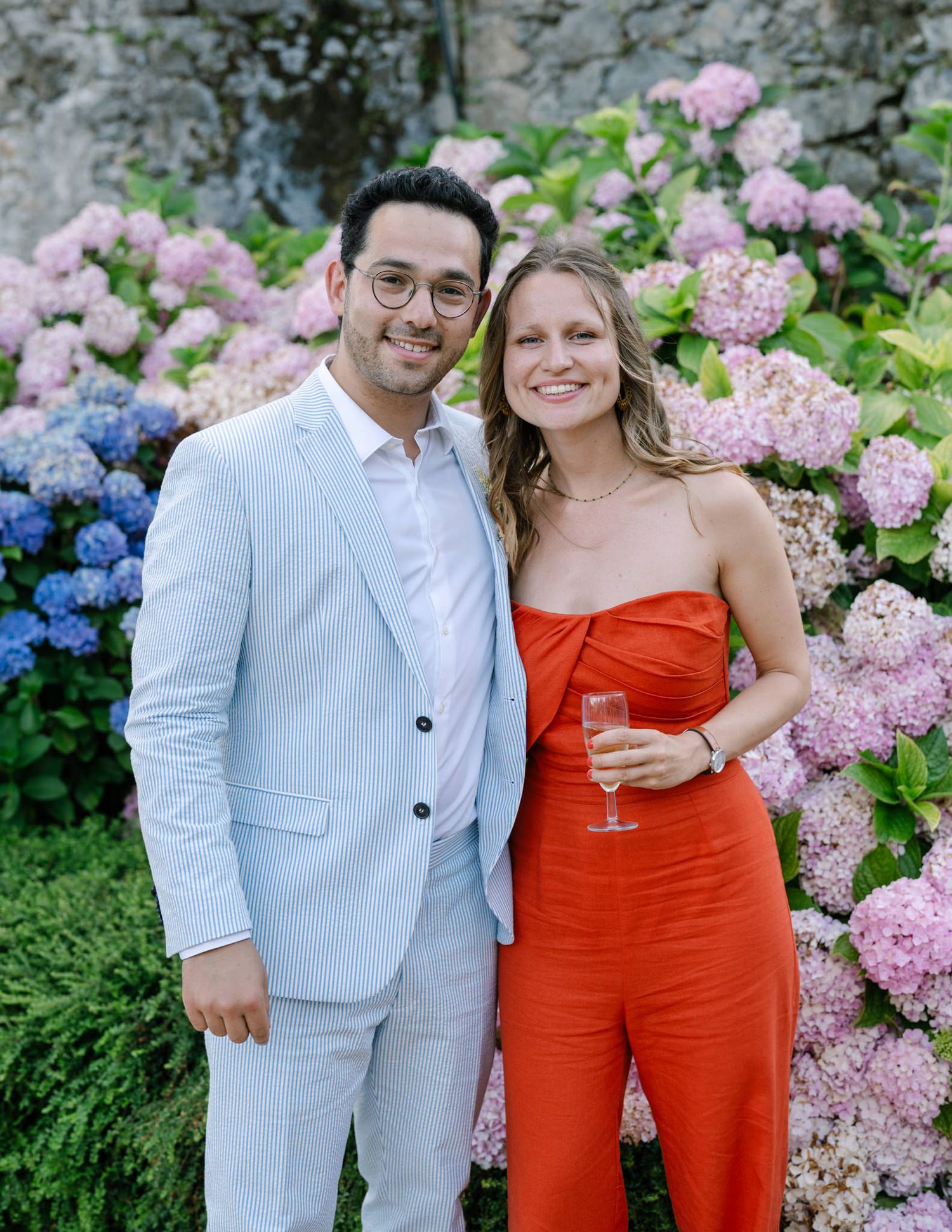 Couple poses among blooming pink and blue hydrangea bushes in a garden setting.