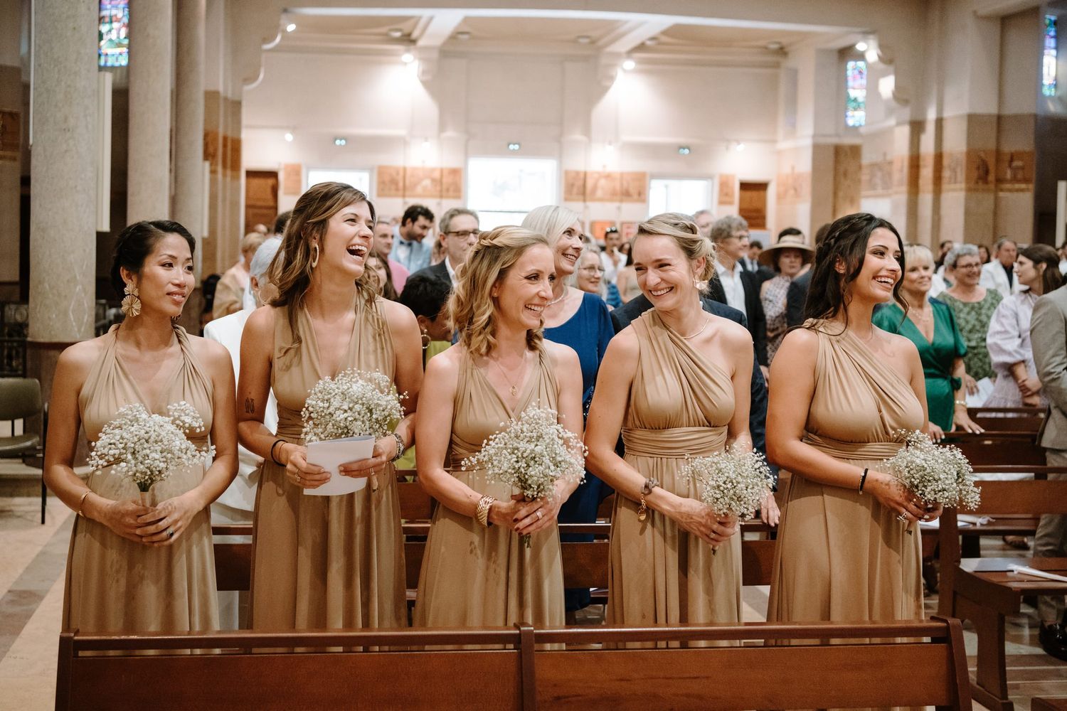 Row of bridesmaids in champagne-colored dresses holding bouquets during church ceremony.